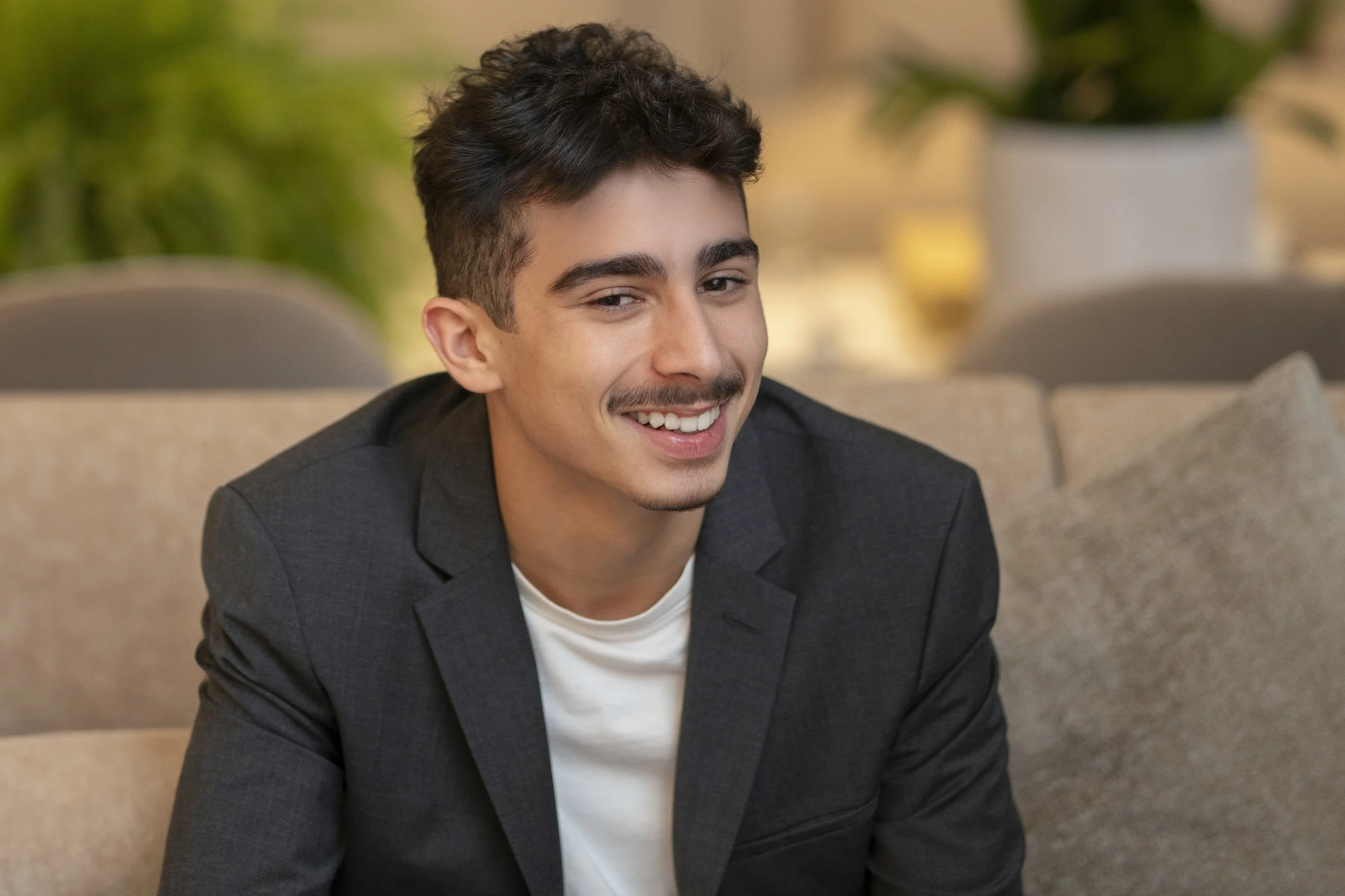 A young man with short dark hair and light facial hair, wearing a black blazer and white shirt, smiling while sitting on a beige couch.