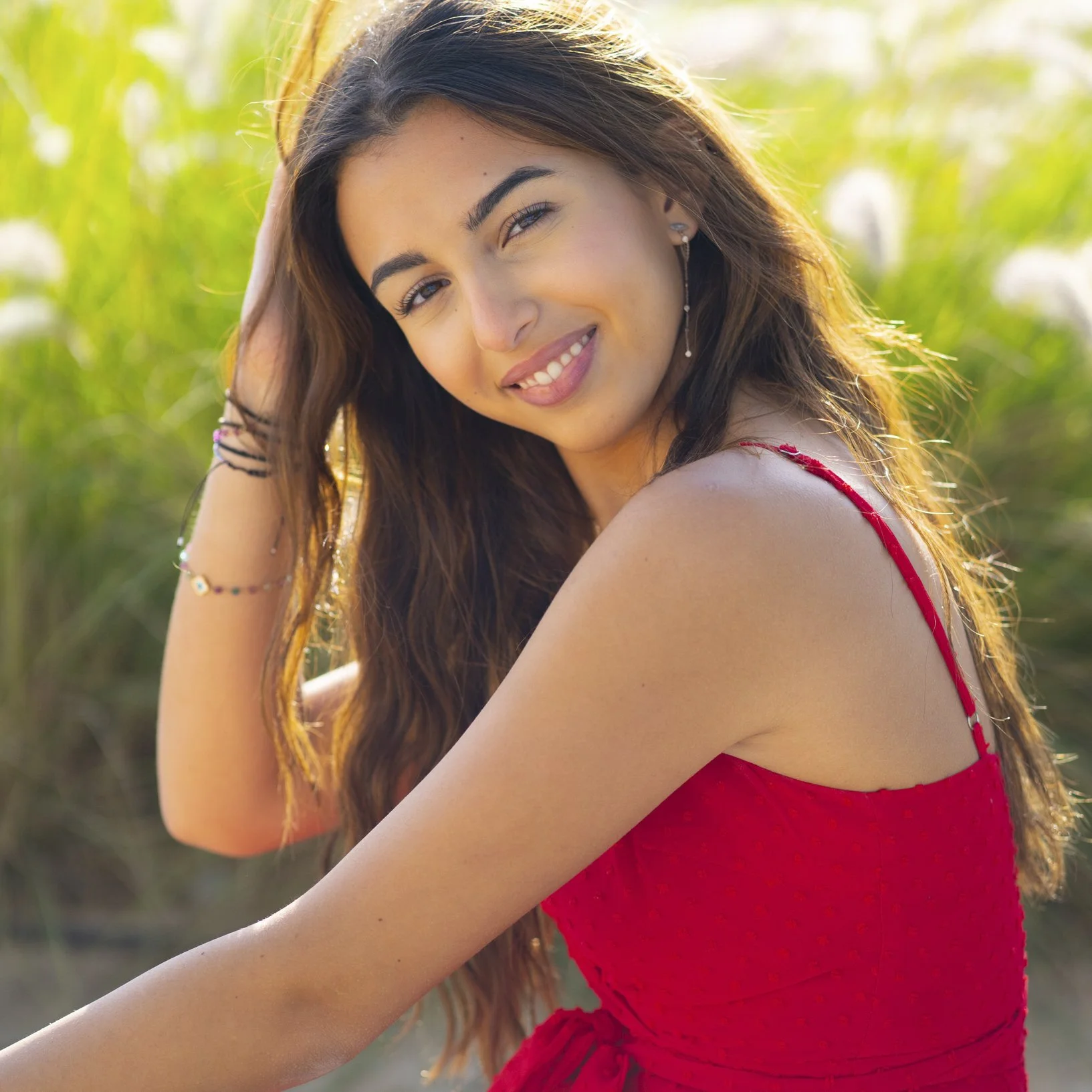 A young woman with long, wavy brown hair, wearing a red spaghetti strap dress, smiling and touching her hair, outdoors in a grassy area with sunlight.