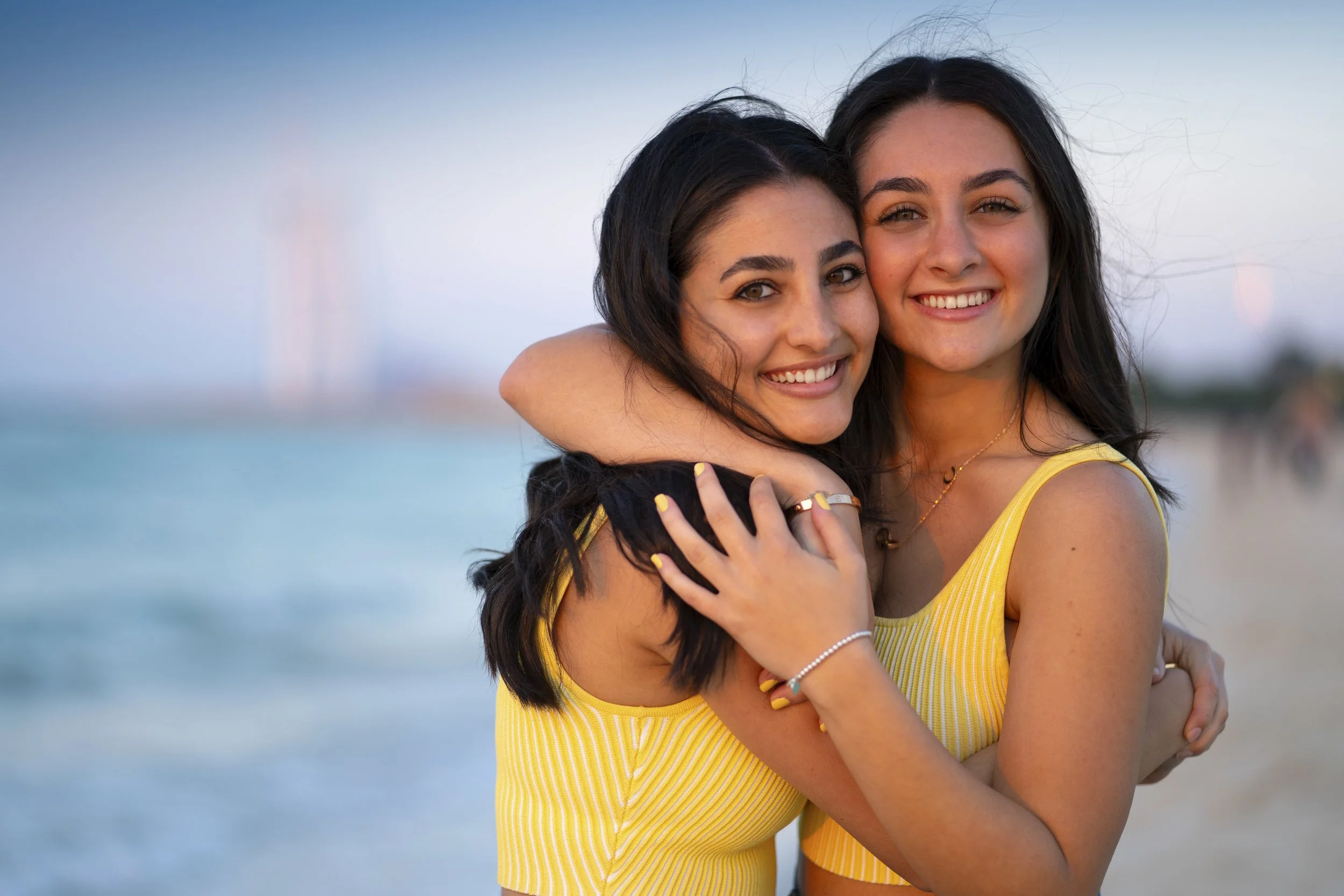 Two young women smiling and hugging on the beach at sunset, wearing yellow striped tops.
