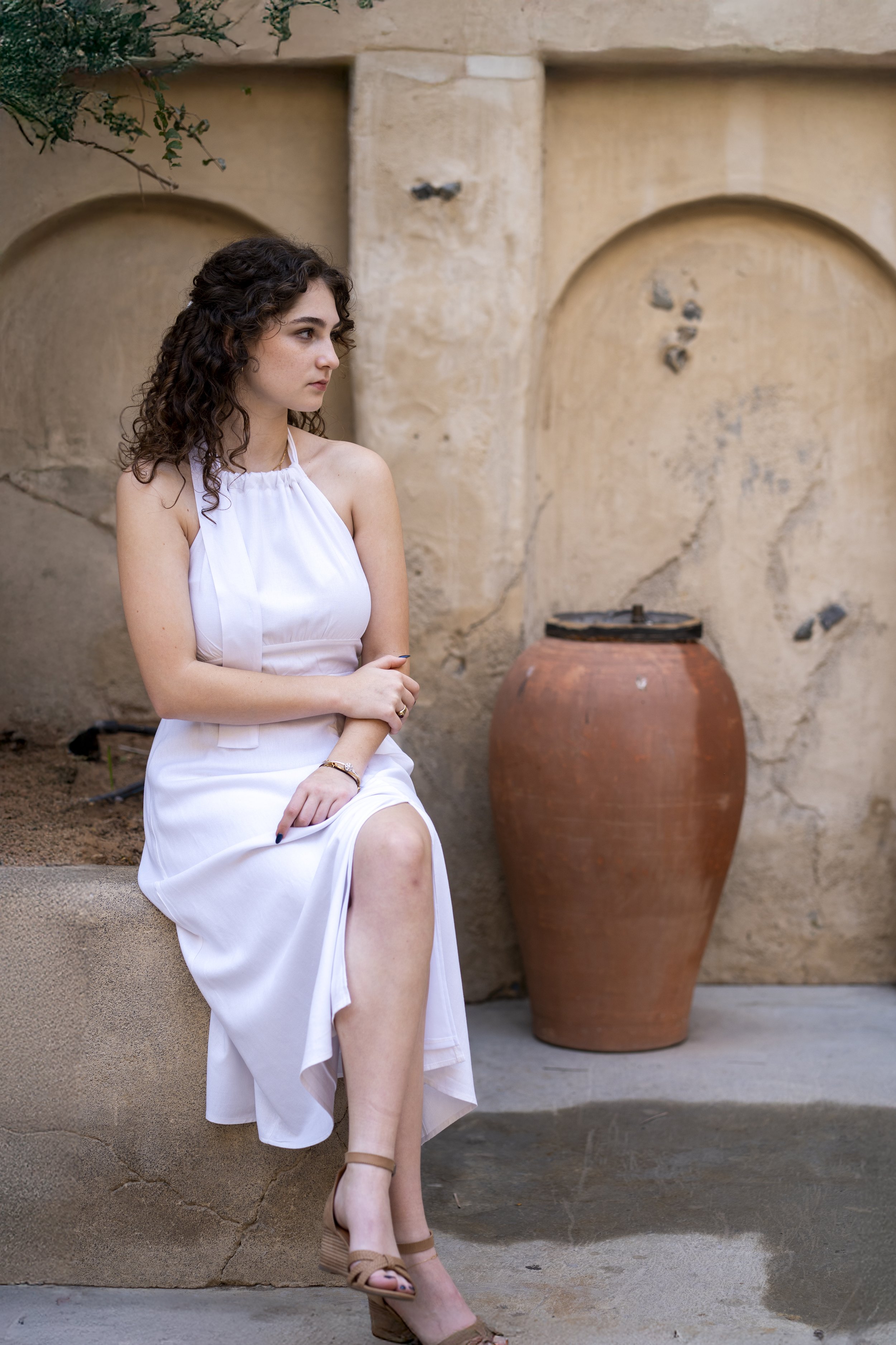 A young woman with curly brown hair wearing a white dress and beige heels sitting on a stone ledge outside, with a large clay pot and a textured beige wall in the background.