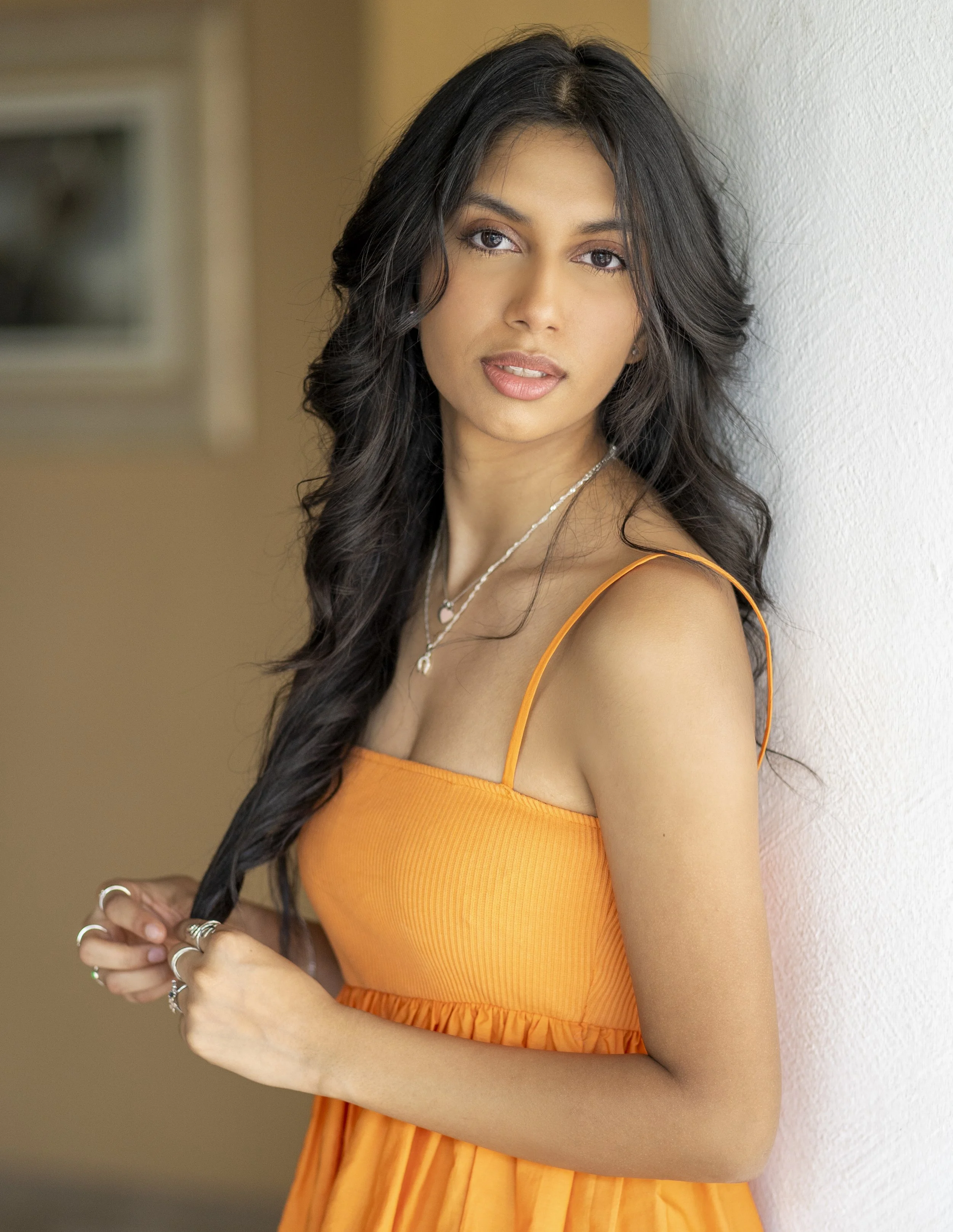 A woman with long dark wavy hair wearing an orange sleeveless dress, standing indoors against a white wall, looking at the camera.
