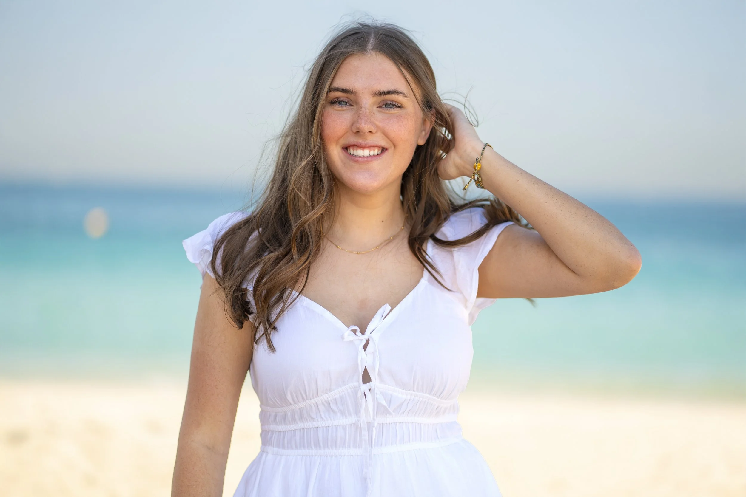 A young woman with long wavy hair smiling at the camera on a beach, wearing a white dress, with the ocean in the background.