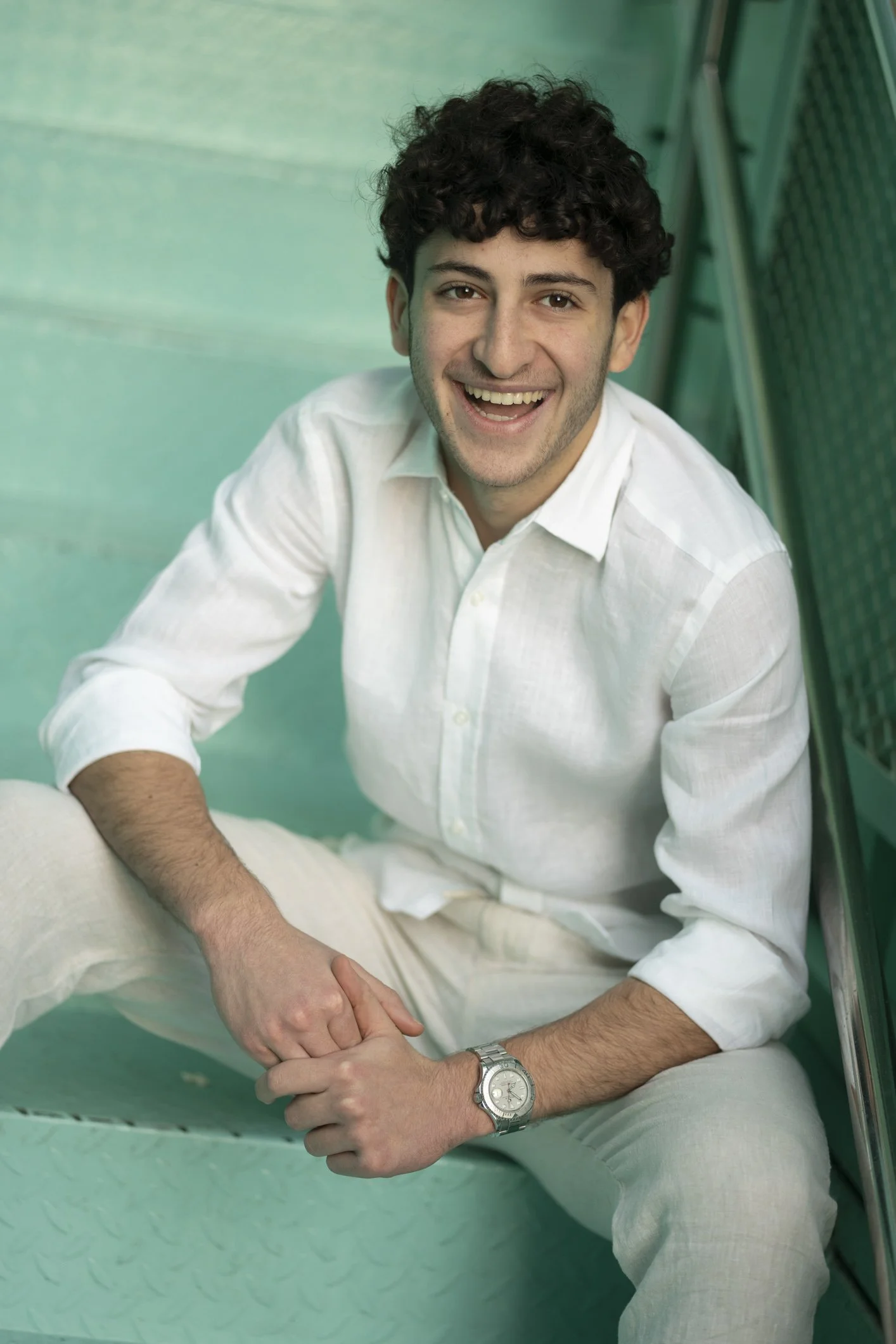 A young man with dark curly hair smiling and sitting on a mint green staircase, wearing a white button-up shirt and a silver wristwatch.