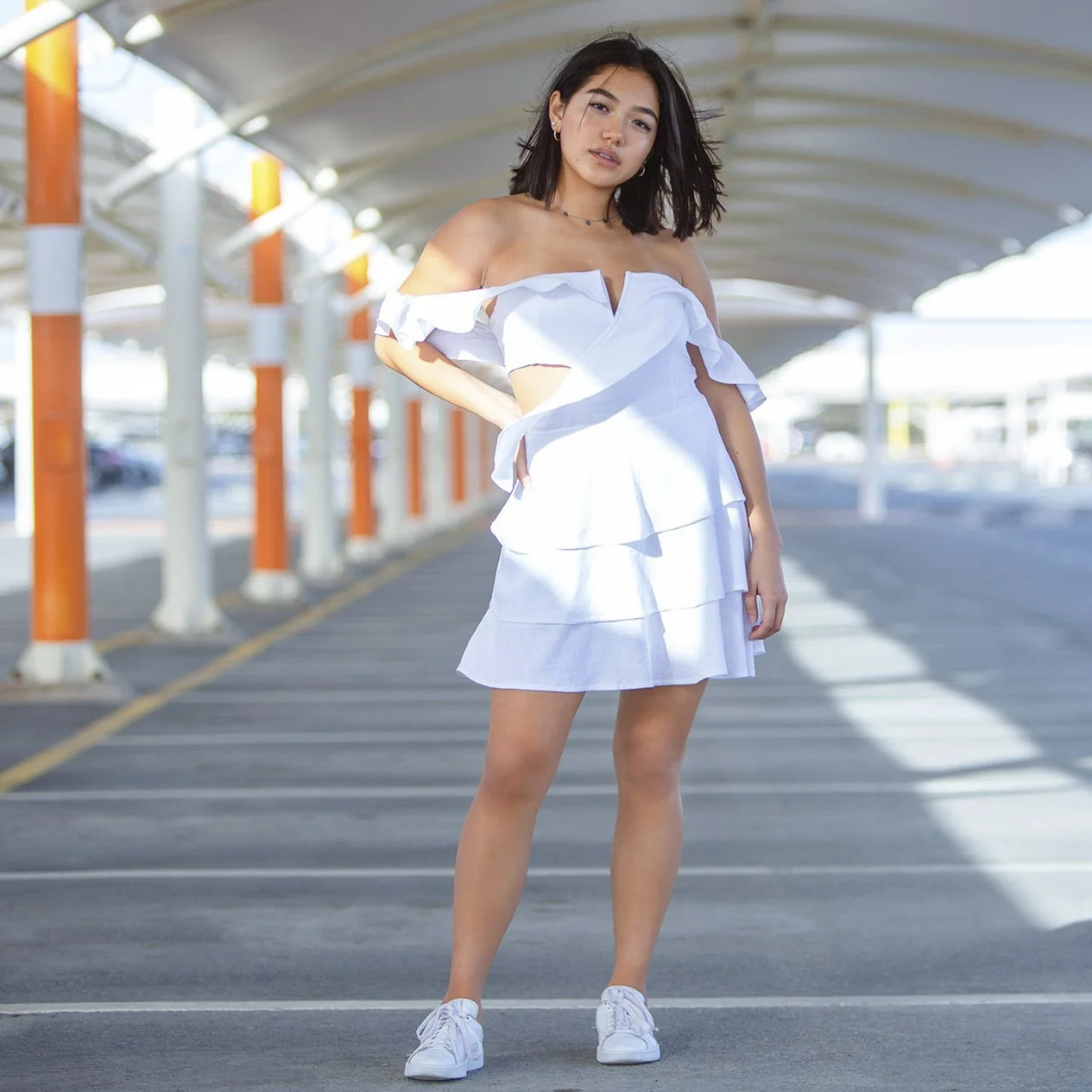 A young woman wearing a white off-shoulder ruffled dress and white sneakers standing in an open-air parking garage.