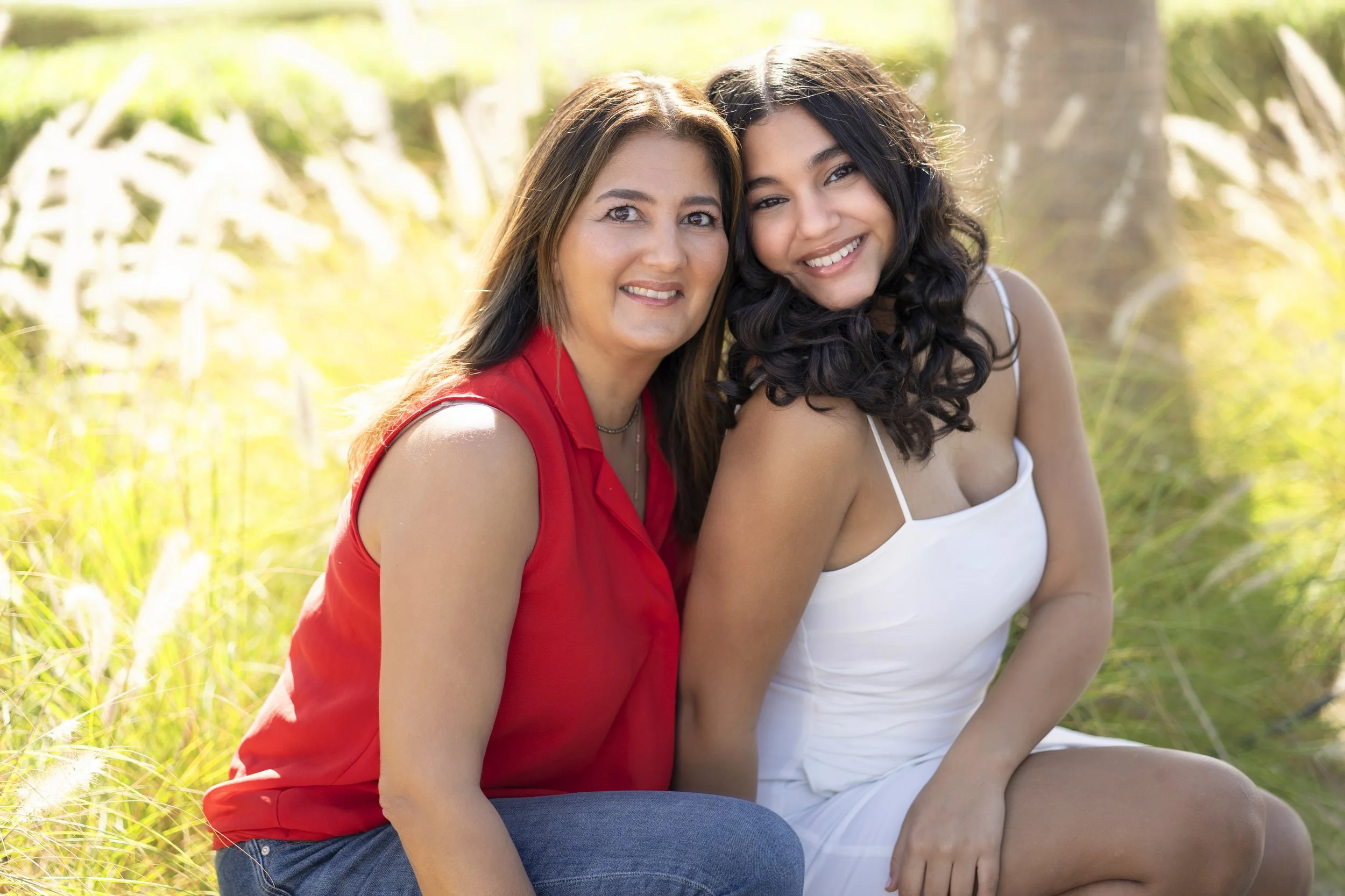 Two women sitting outdoors in a grassy area, smiling at the camera. The woman on the left has long brown hair, wearing a red sleeveless top, and the woman on the right has long dark curly hair, wearing a white dress.