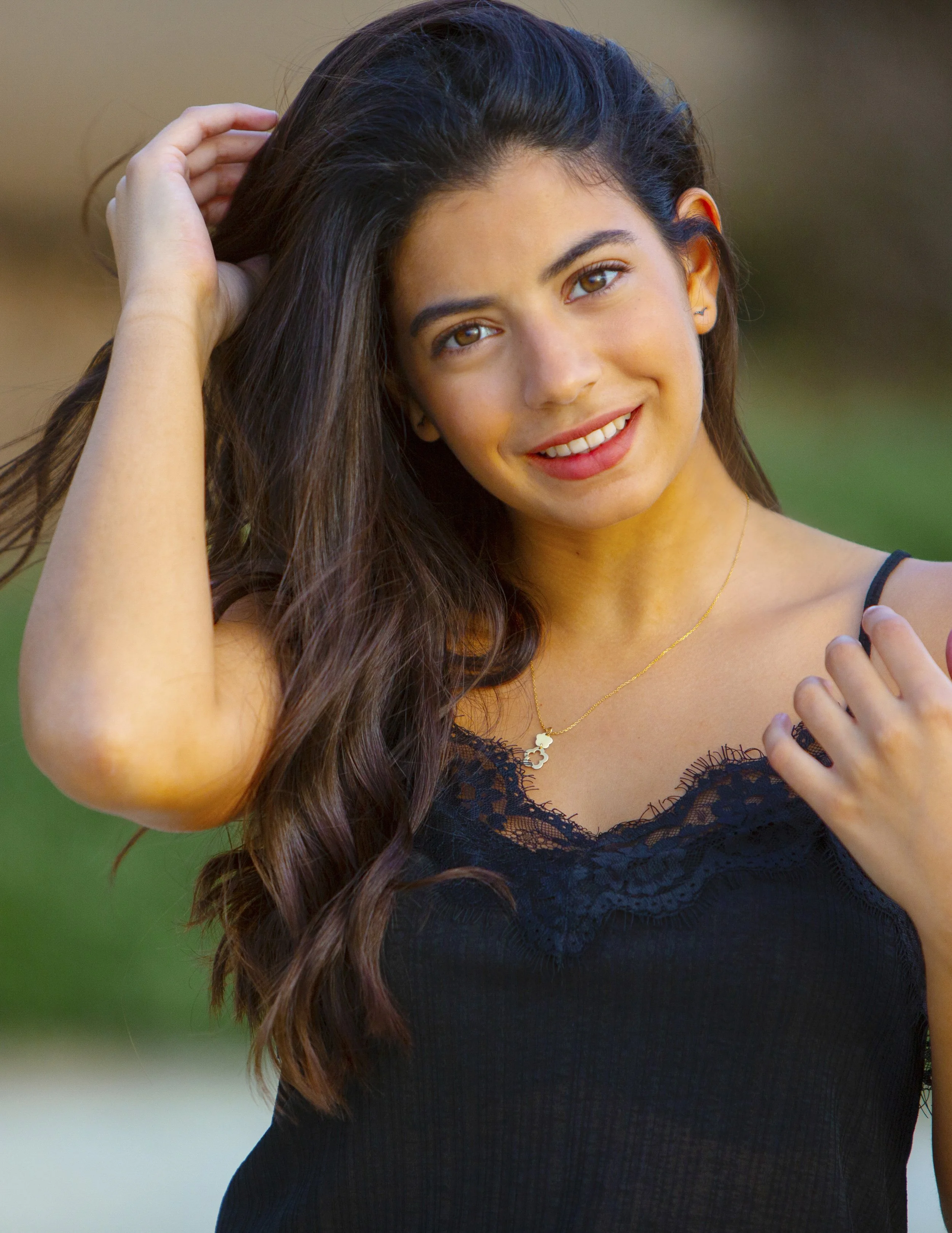 A young woman with long, wavy brown hair, smiling, wearing a black lace-trimmed top, gold necklace, and small earrings, outdoors with a blurred background.