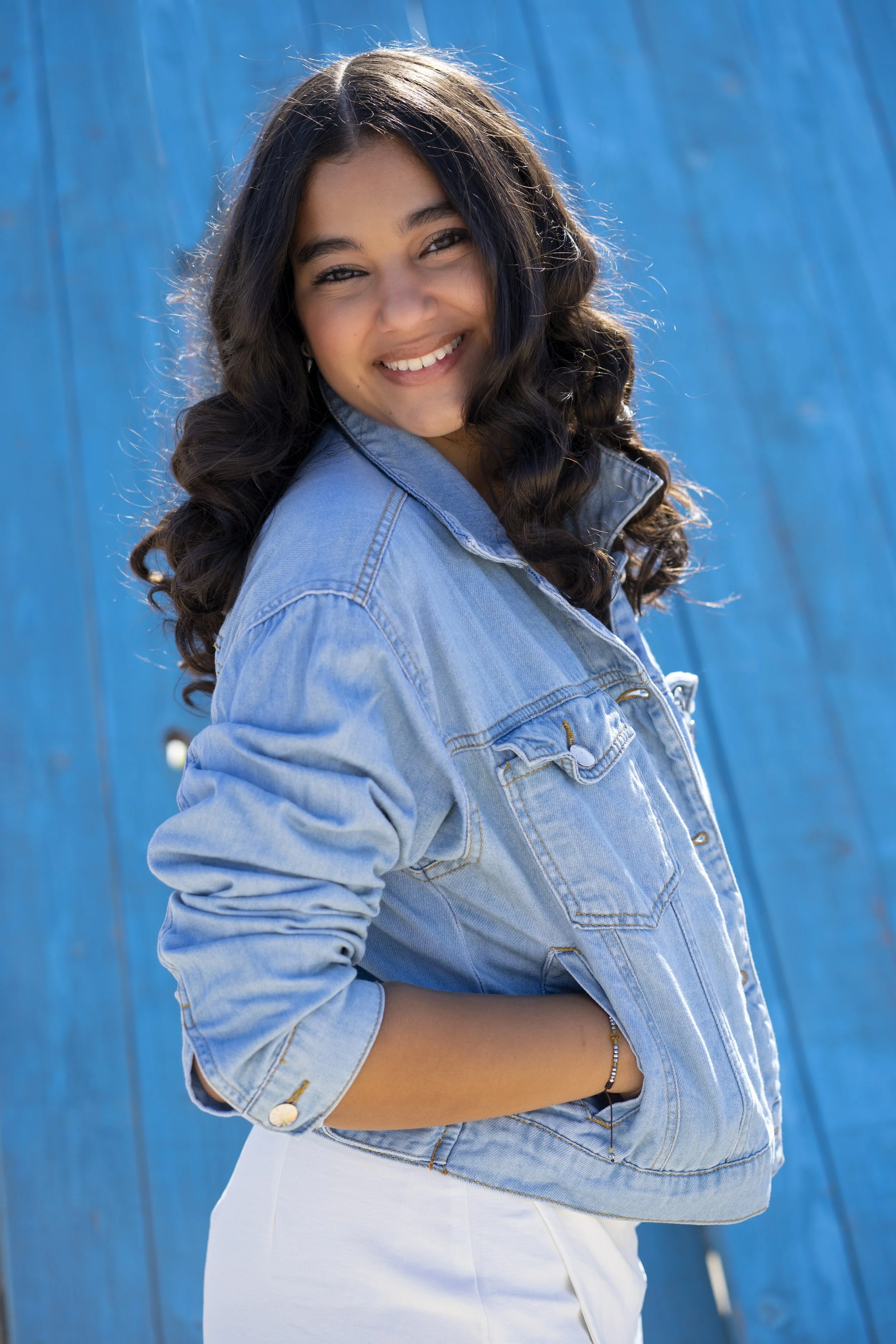 Young woman with long curly dark hair wearing a light blue denim jacket and white pants, smiling outdoors against a blue background.