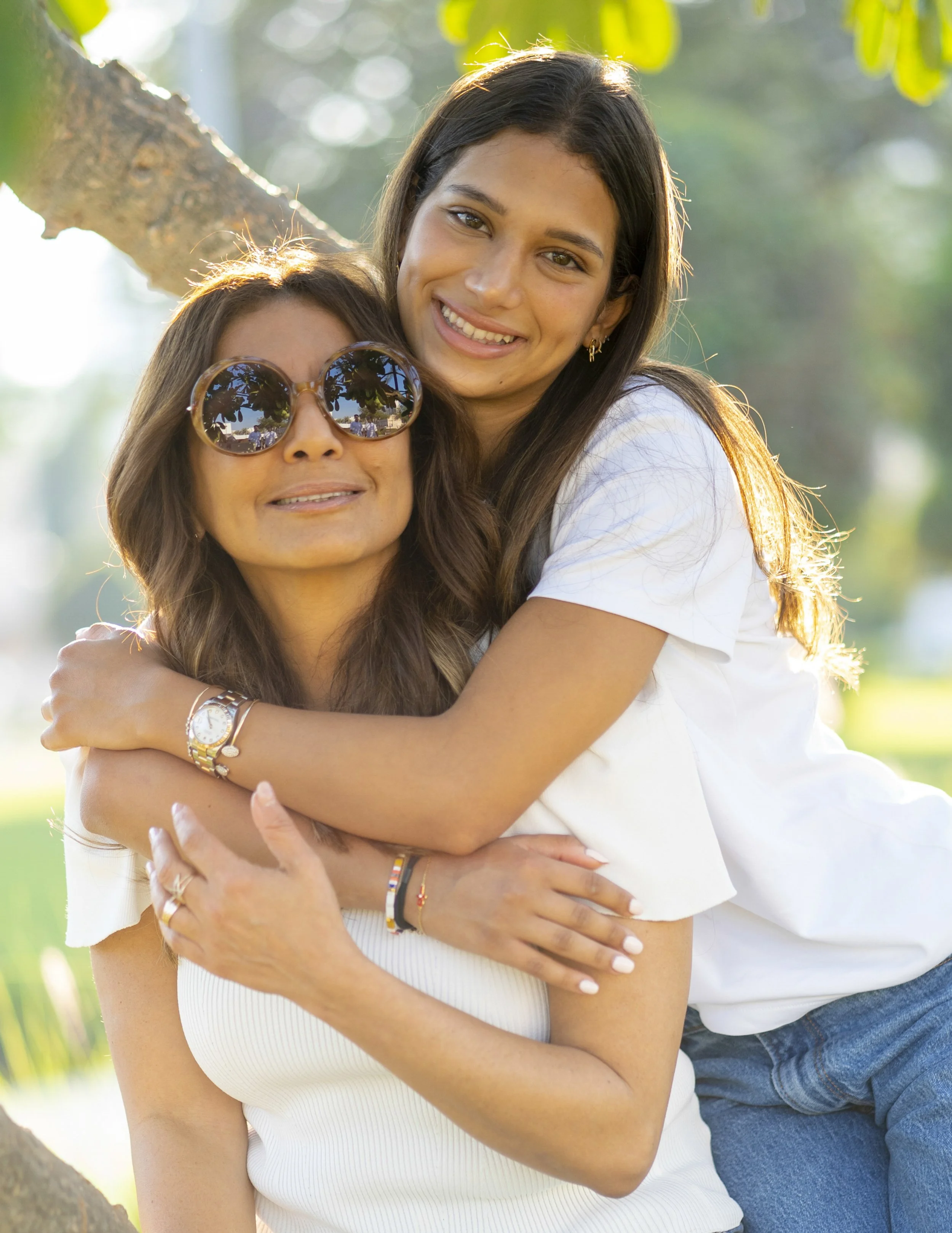 Two women hugging outdoors on a sunny day, one wearing sunglasses and the other with long dark hair, both smiling.