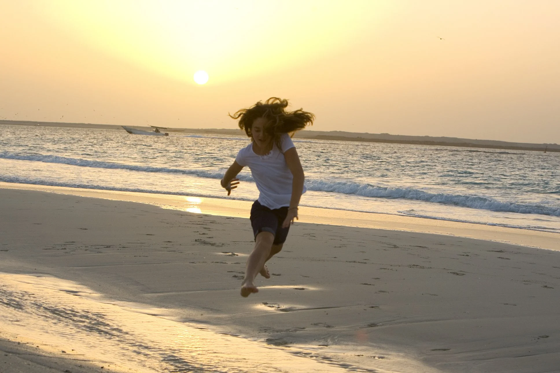 A person running barefoot on the beach during sunset, with the ocean and a boat in the background.