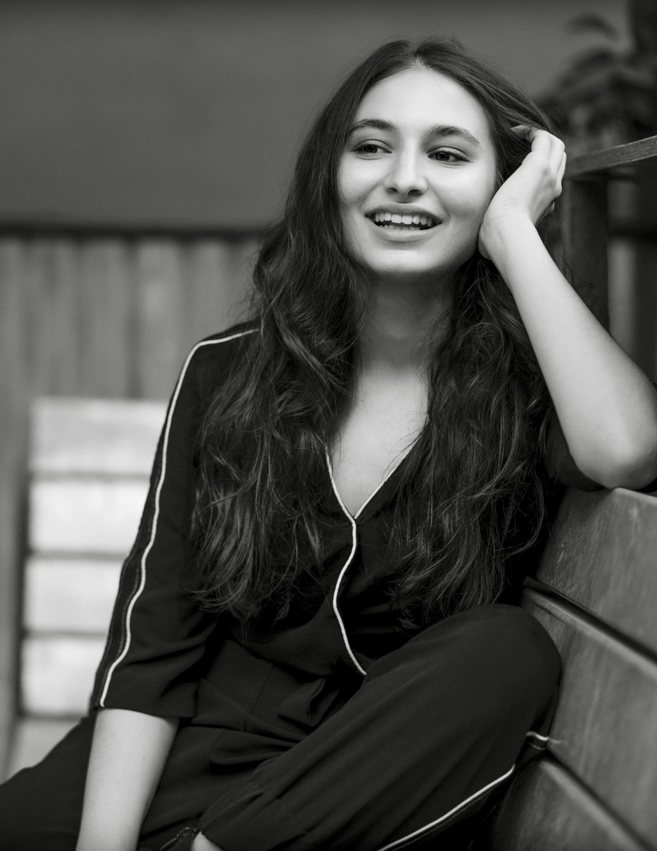 A young woman with long wavy hair, smiling, sitting on a bench with her hand resting on her head in a monochrome photo.