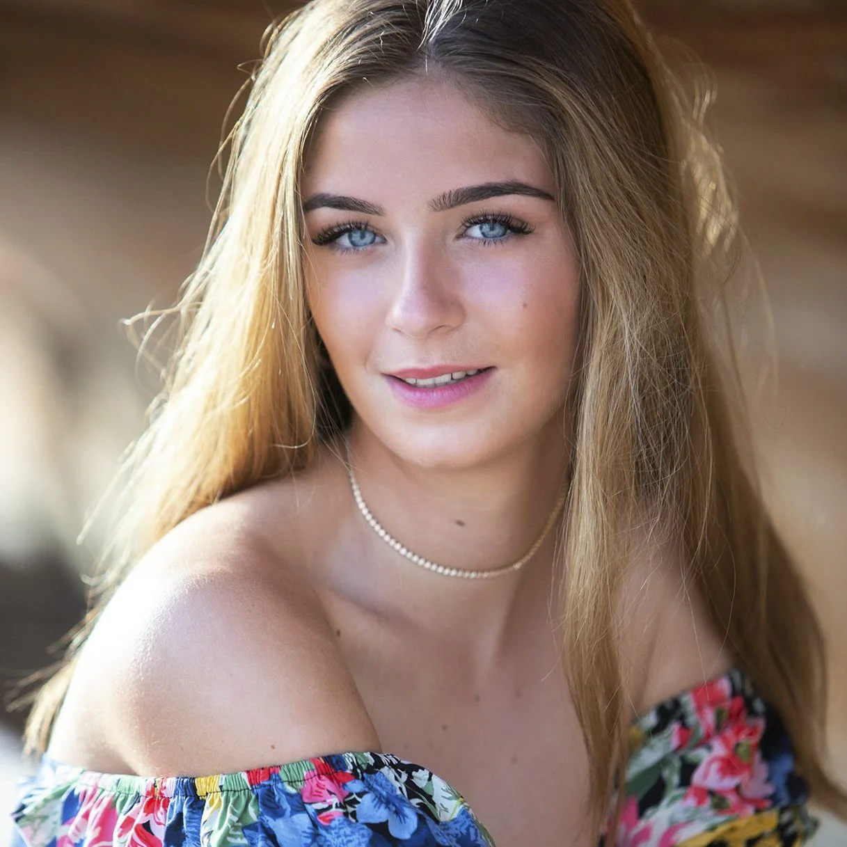 A young woman with long, light brown hair and bright blue eyes. She is smiling softly, wearing a colorful off-the-shoulder top with a floral pattern, and a delicate pearl necklace. The background is blurred with warm lighting.
