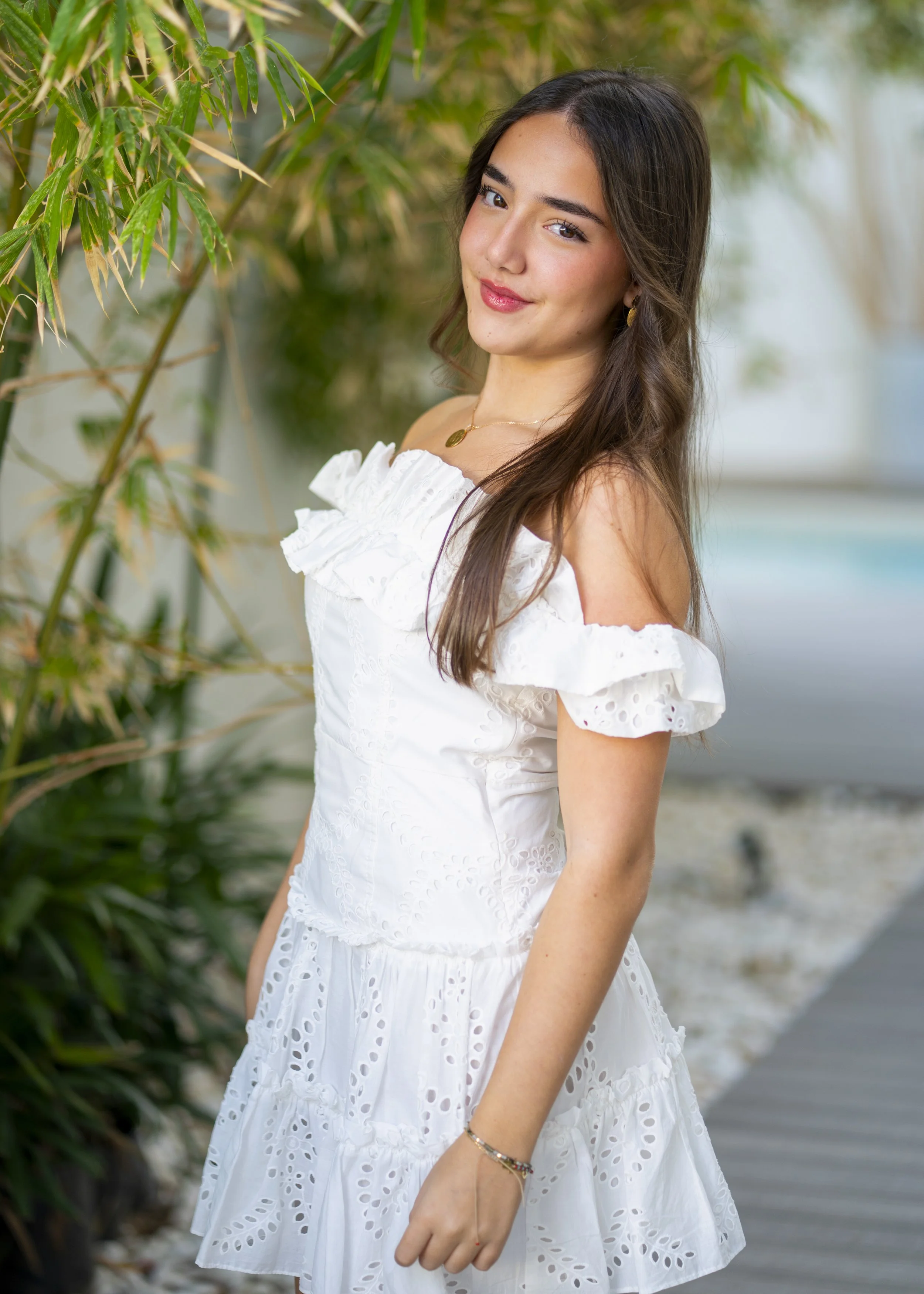 A young woman with long brown hair, wearing a white off-shoulder dress with eyelet details, standing outdoors near green foliage and a pathway.