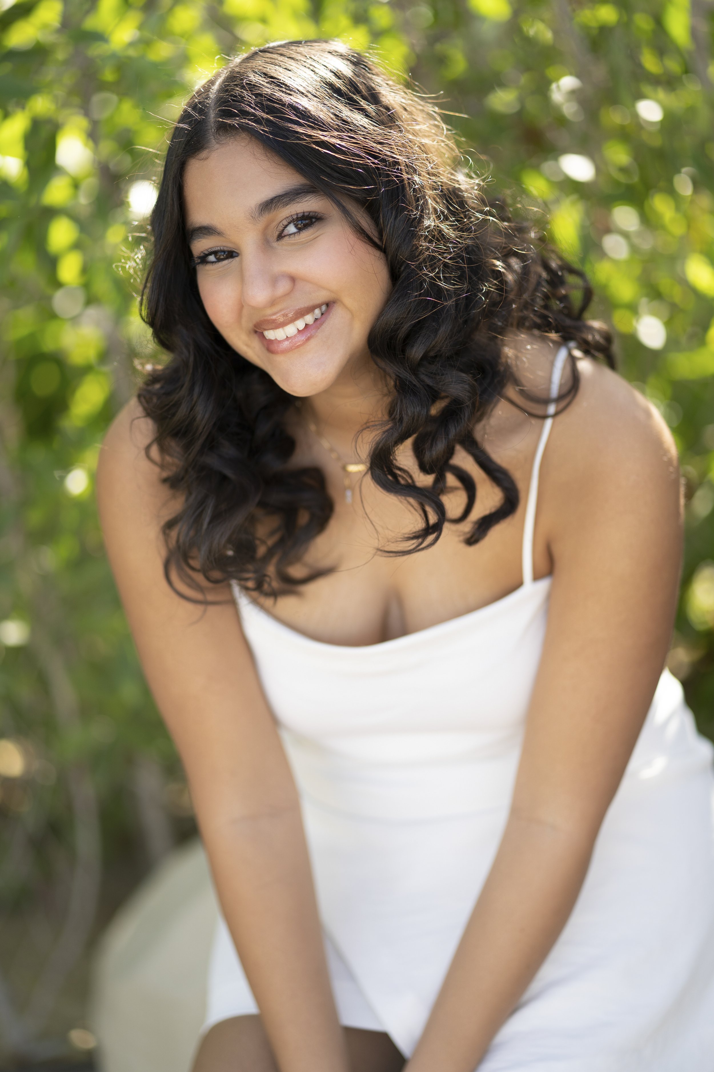A young woman with long, dark, curly hair smiling outdoors with green foliage in the background, wearing a white dress with spaghetti straps.