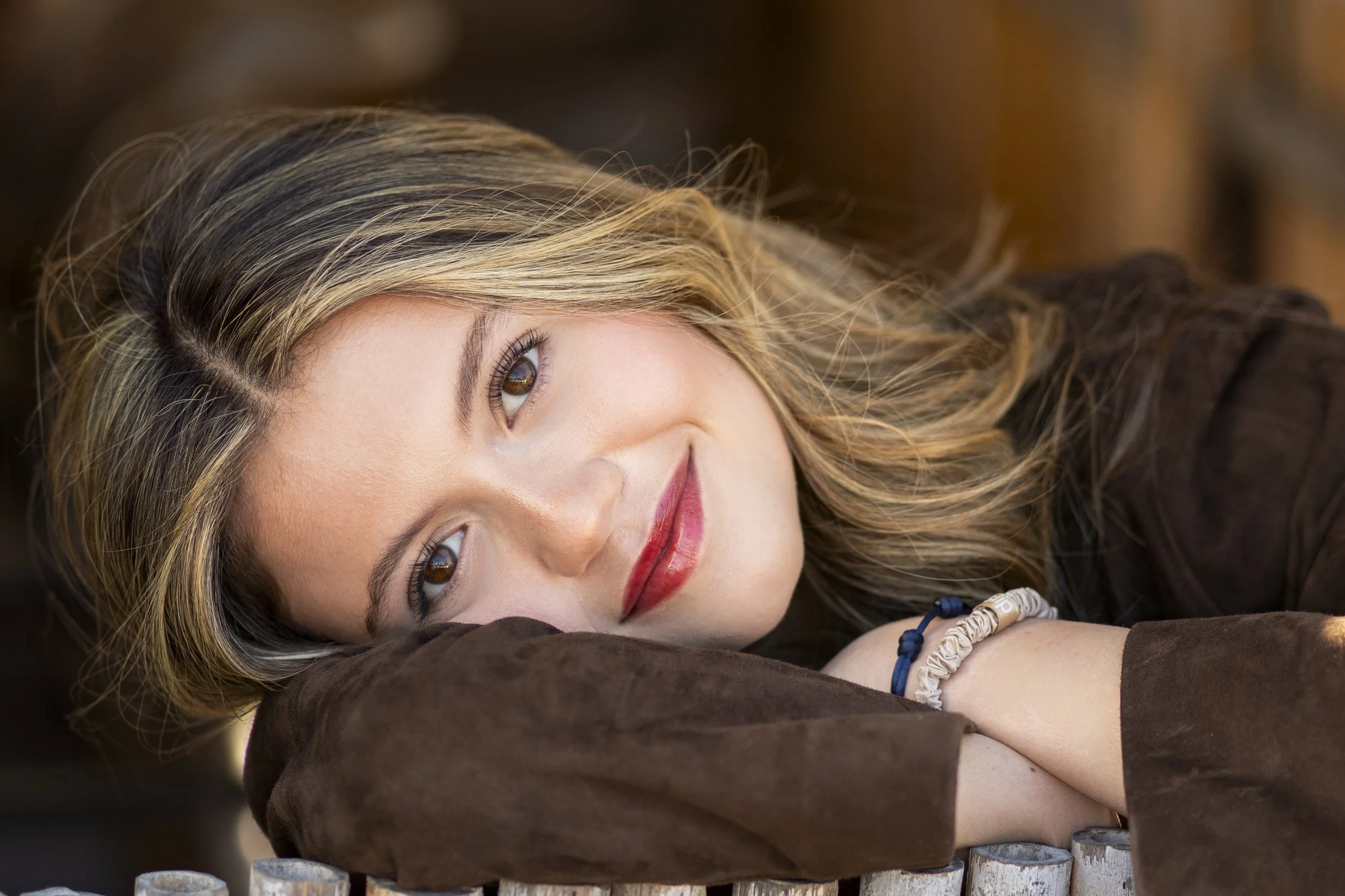 A woman with blonde and brown hair lying on her side on a brown pillow, resting her head on her arm, and looking at the camera with a soft smile.