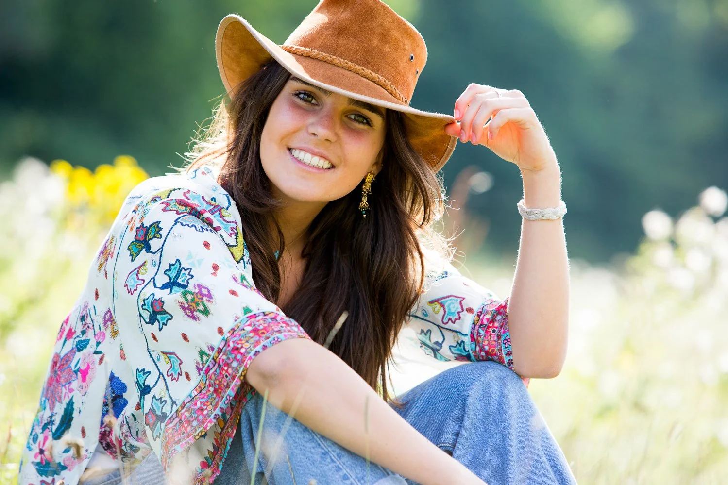 A woman in a colorful bohemian blouse and blue jeans, smiling and sitting outdoors in a grassy field, wearing a brown wide-brimmed hat, with a blurred natural background.