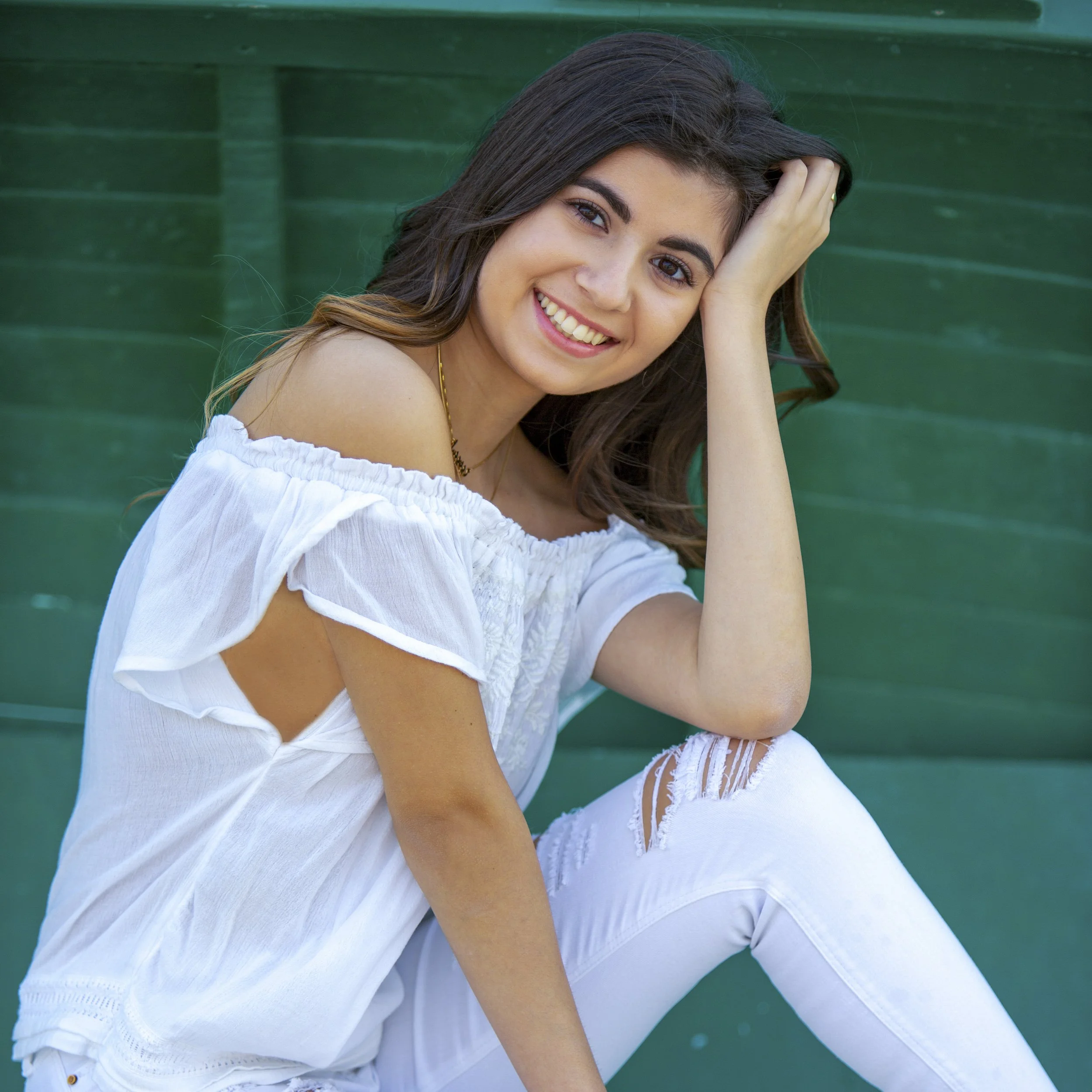 A young woman with long brown hair sitting in front of a green wooden background, smiling, wearing a white off-shoulder top and ripped white jeans.