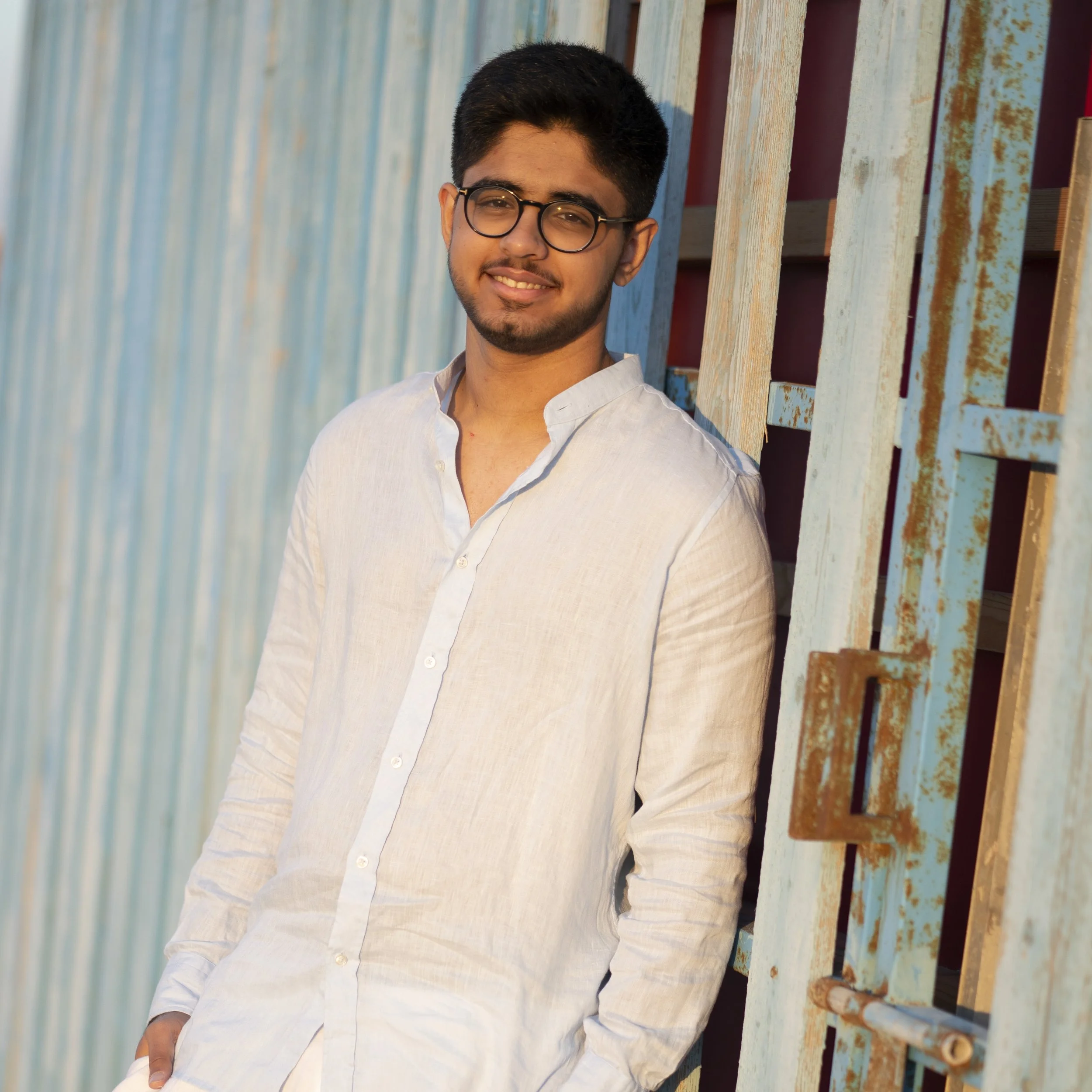 A young man with glasses, a goatee, and dark hair, smiling and wearing a white shirt, standing against a weathered wooden and metal fence.