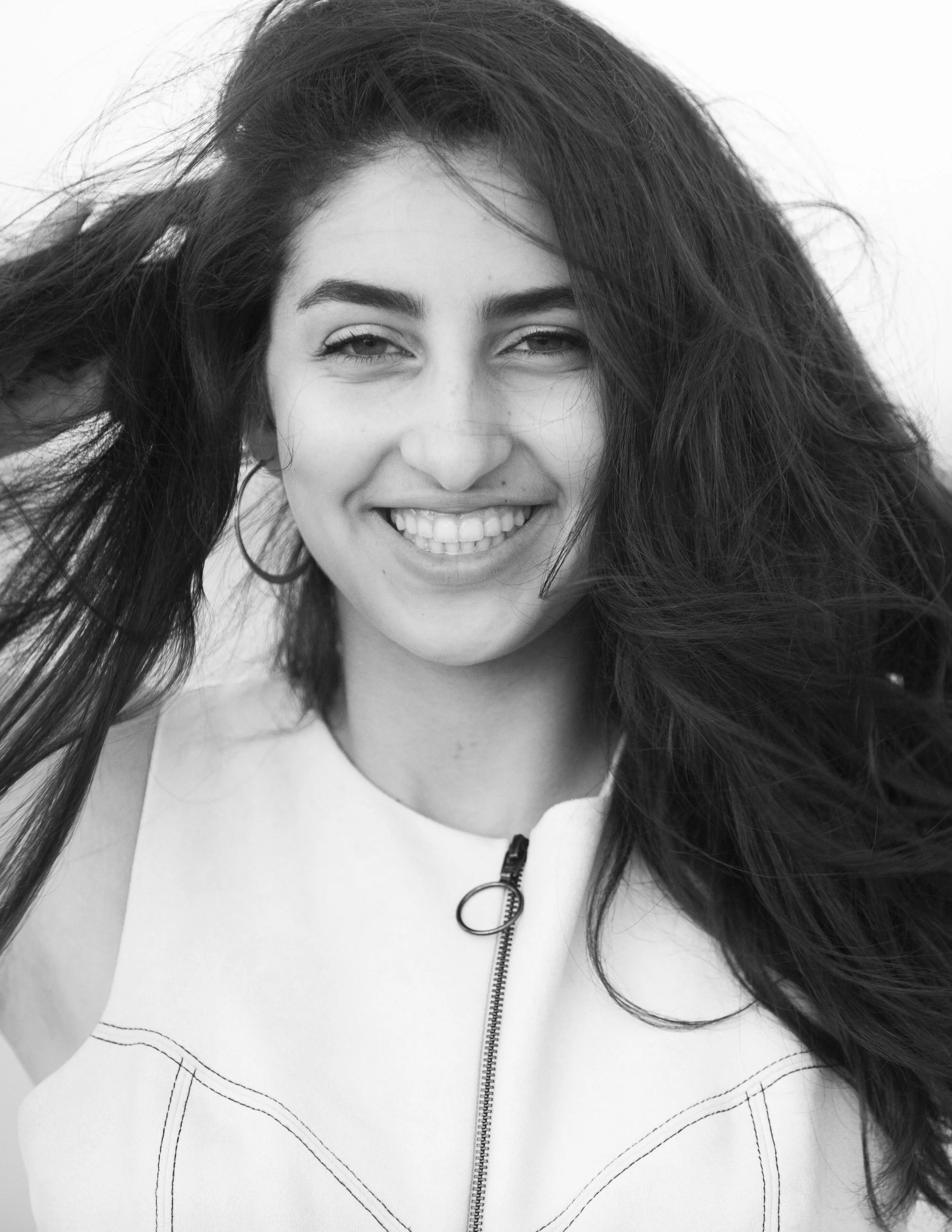 Black and white photo of a young woman smiling, with long hair blowing to the side, wearing a zip-up top.