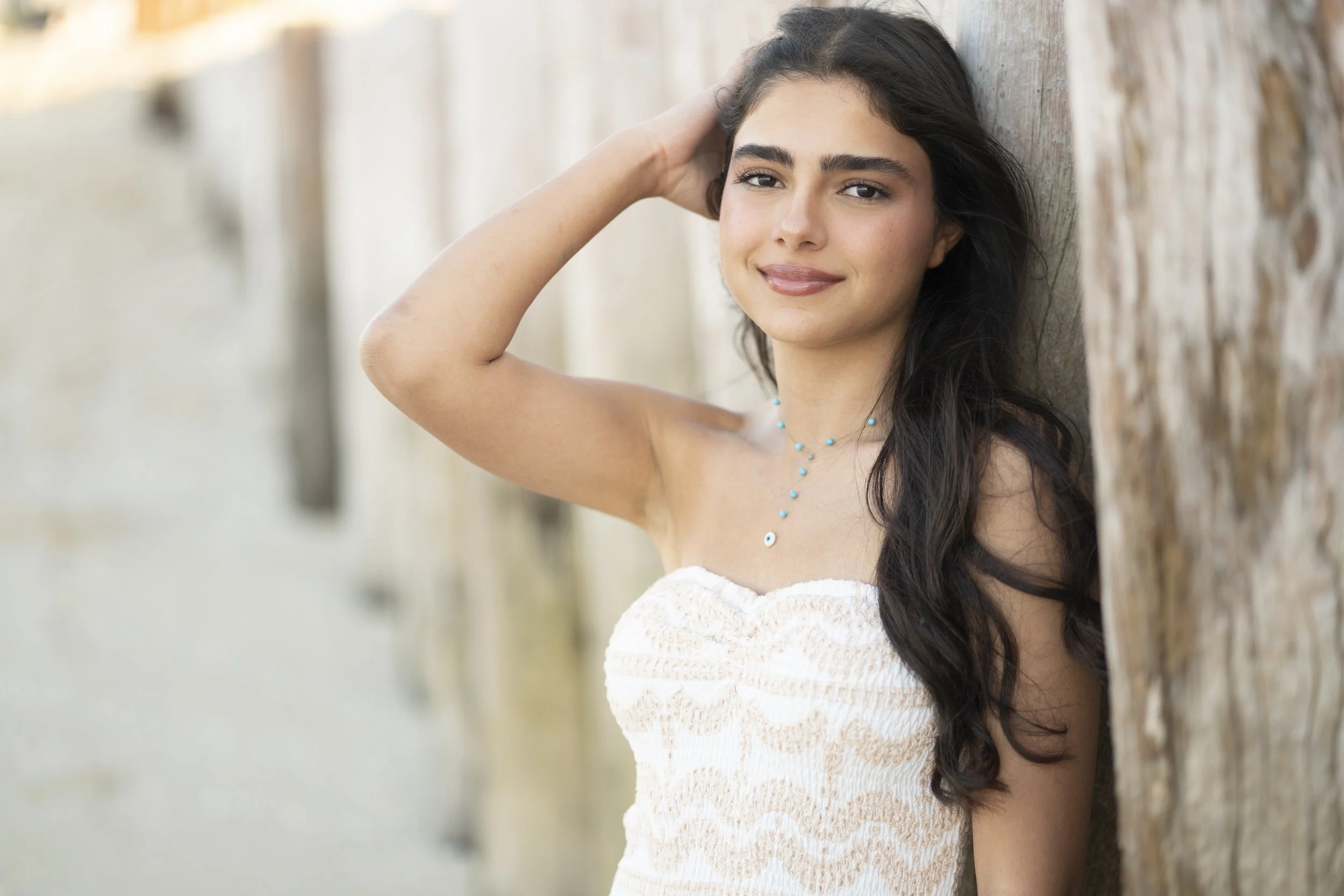 Young woman with long dark hair leaning against a wooden wall, smiling, wearing a white strapless dress and a turquoise beaded necklace.