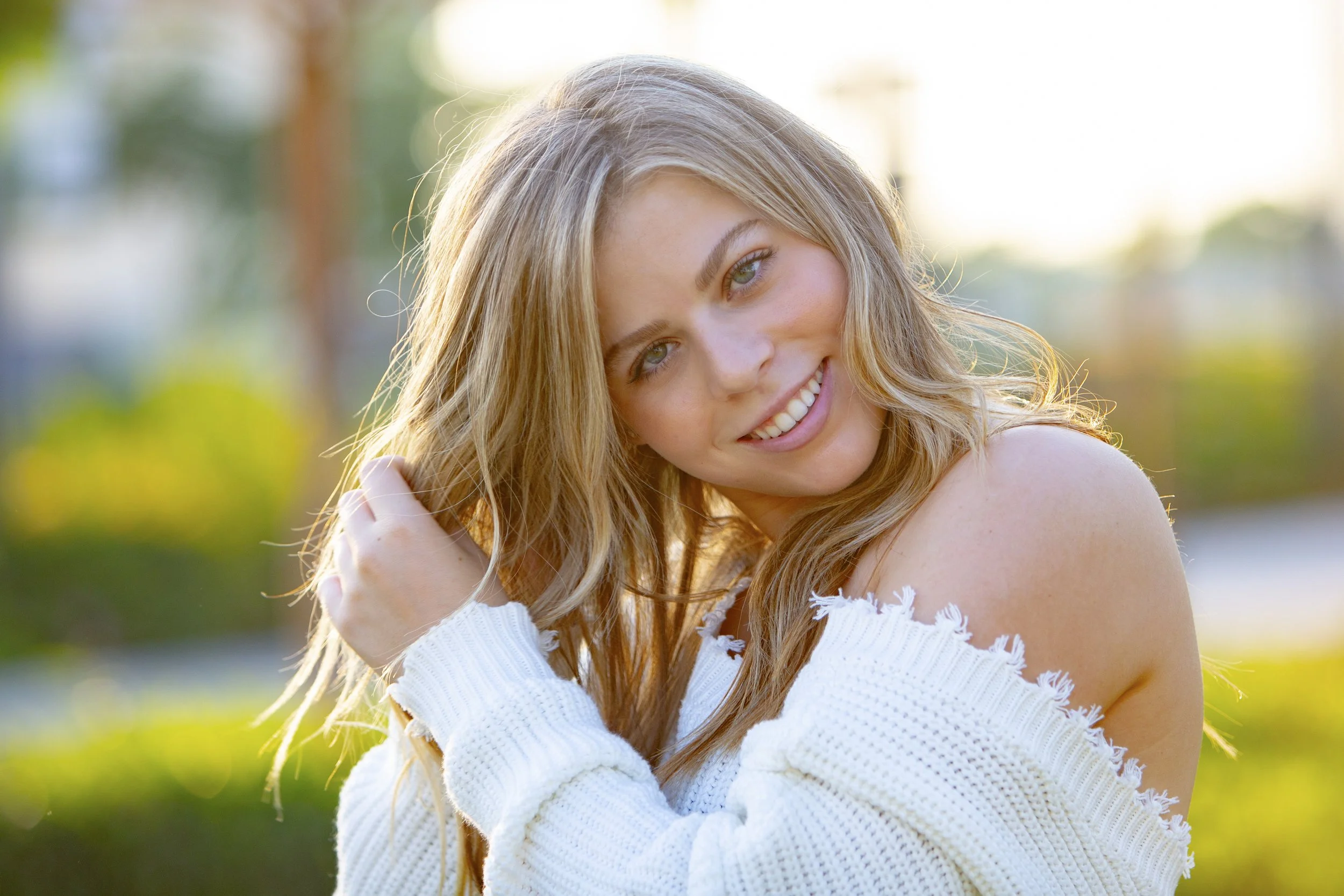 A young woman with blonde hair smiling at the camera outdoors during sunset, wearing a white knitted sweater.