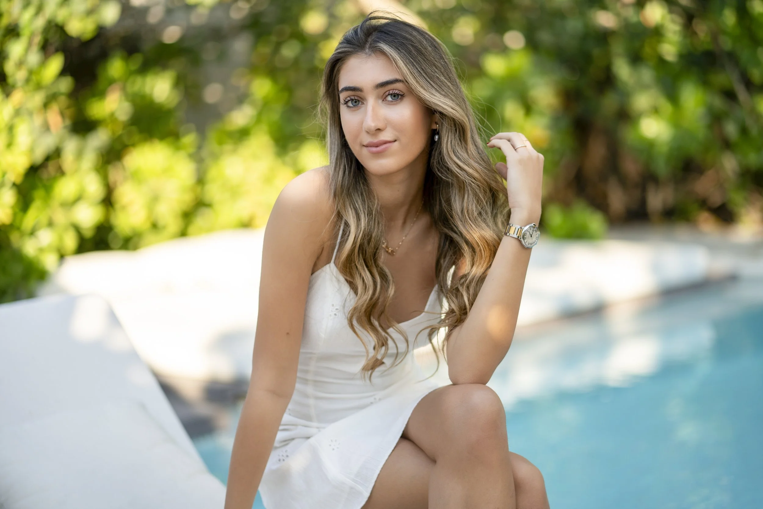 Young woman with wavy hair sitting outdoors by a pool, wearing a white dress and a watch, with greenery in the background.