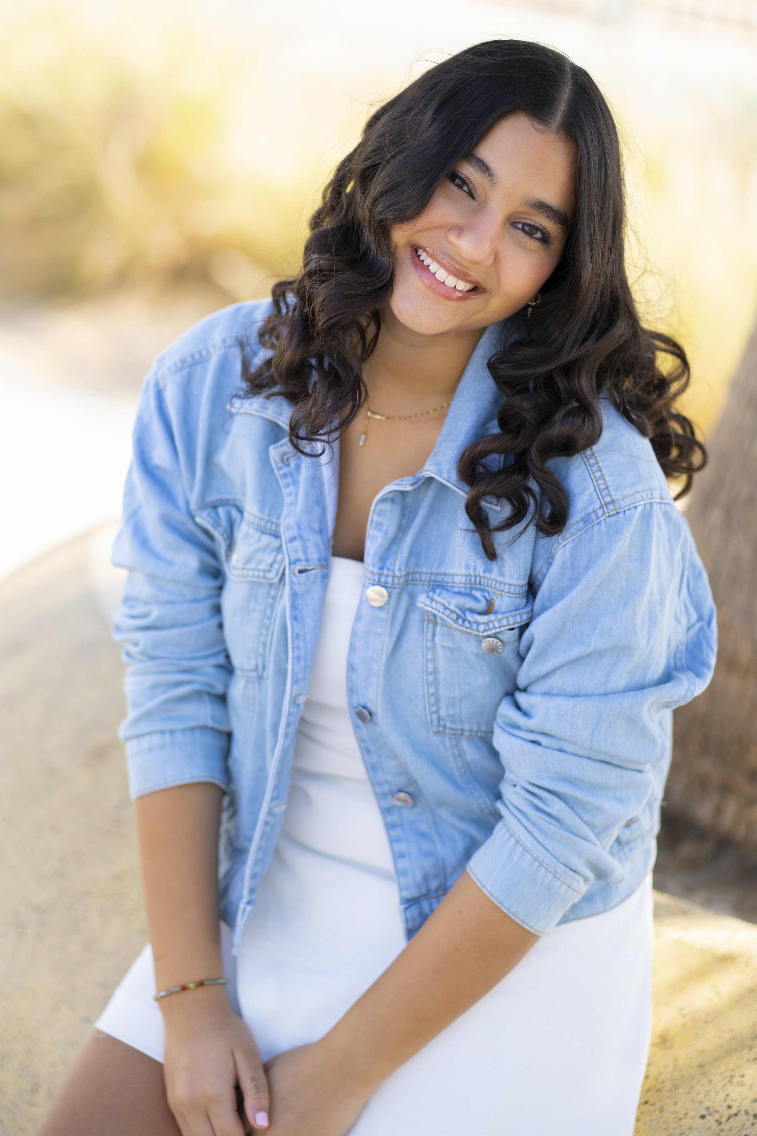A young woman with dark curly hair and tan skin outdoors, smiling and wearing a light blue denim jacket over a white top, with a blurred background of trees and sunlight.