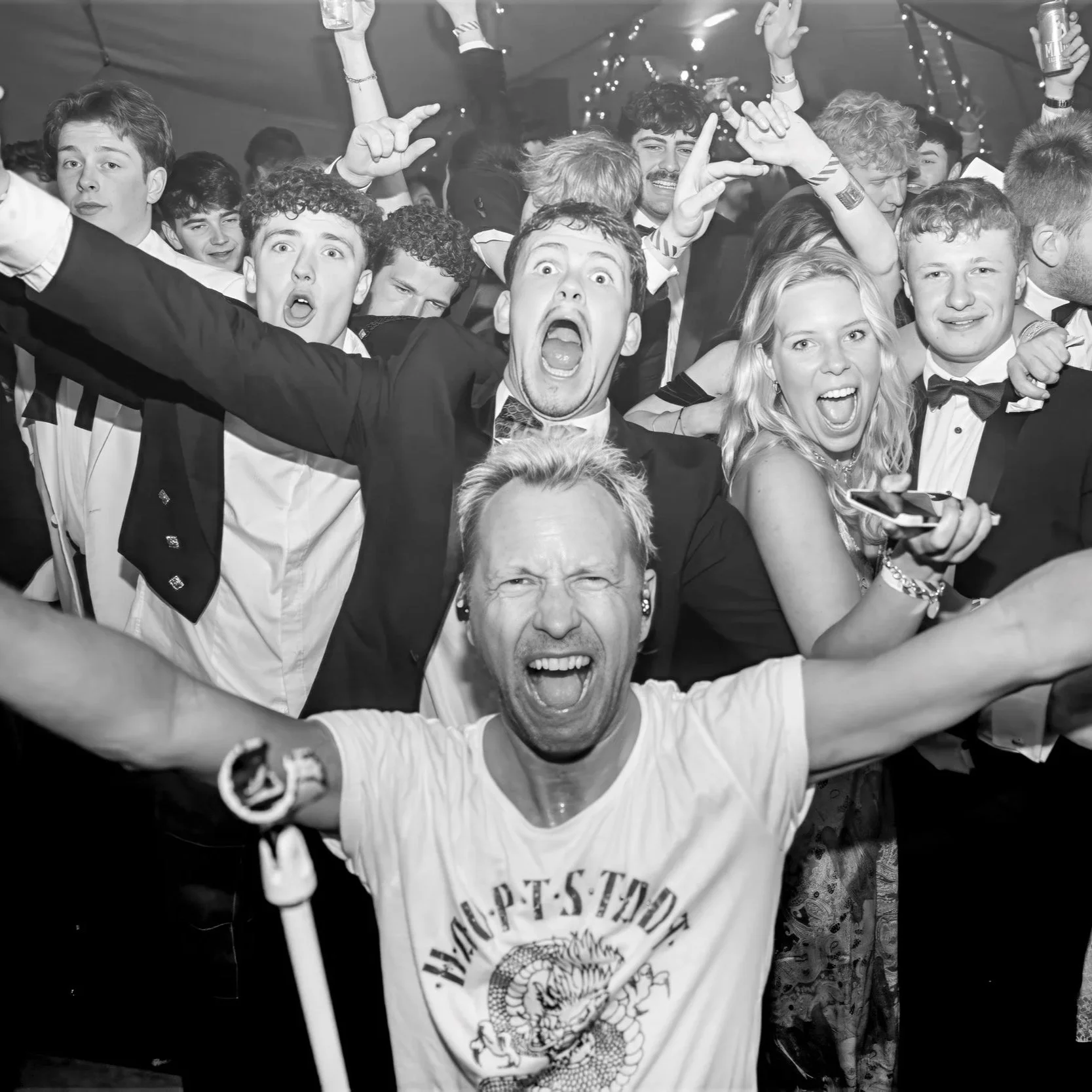 A group of people at a party, smiling and cheering with arms raised, dressed in formal attire, with a man in a casual t-shirt in the foreground.
