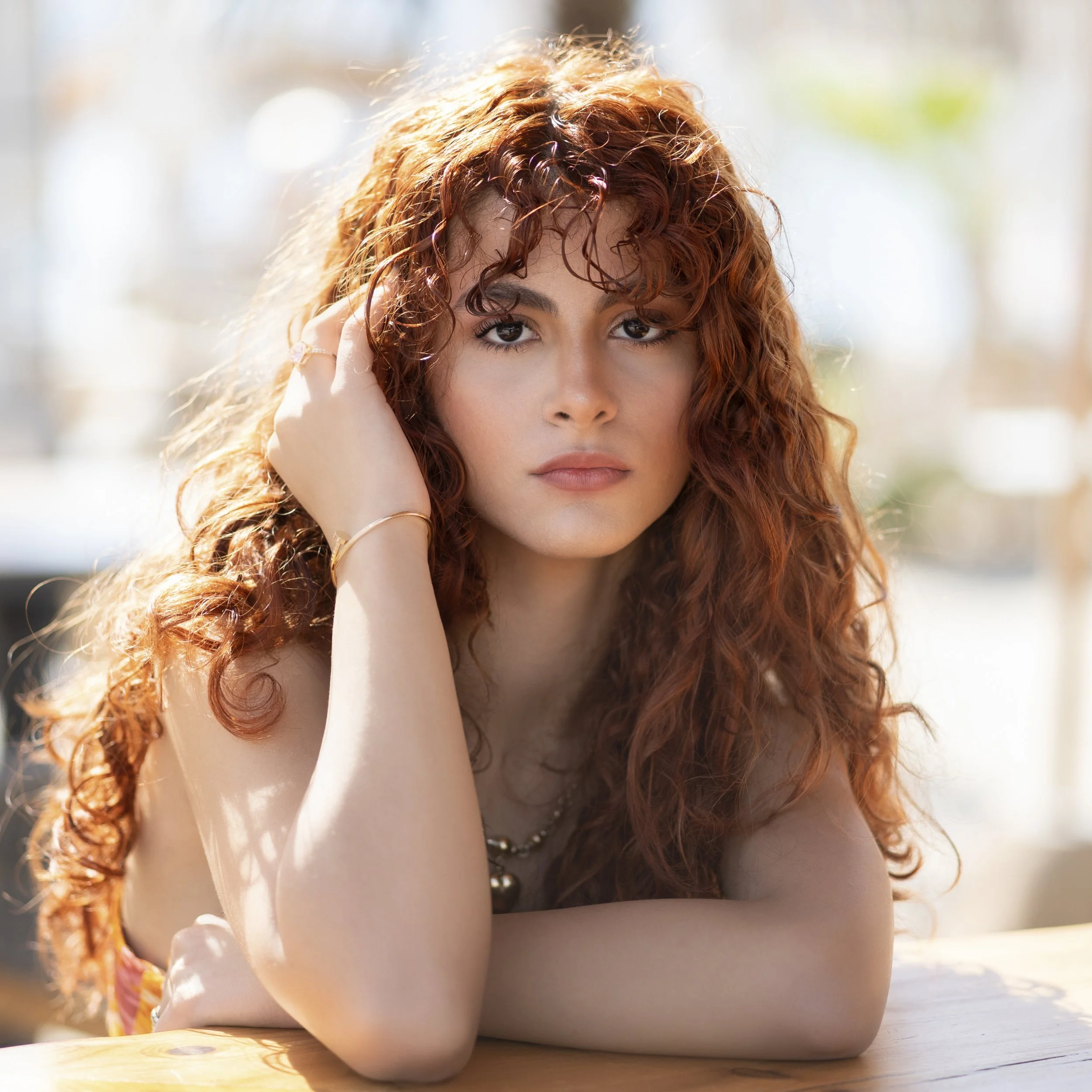 A young woman with long, curly red hair looks directly at the camera, resting her head on her left hand, with an outdoor background and soft lighting.