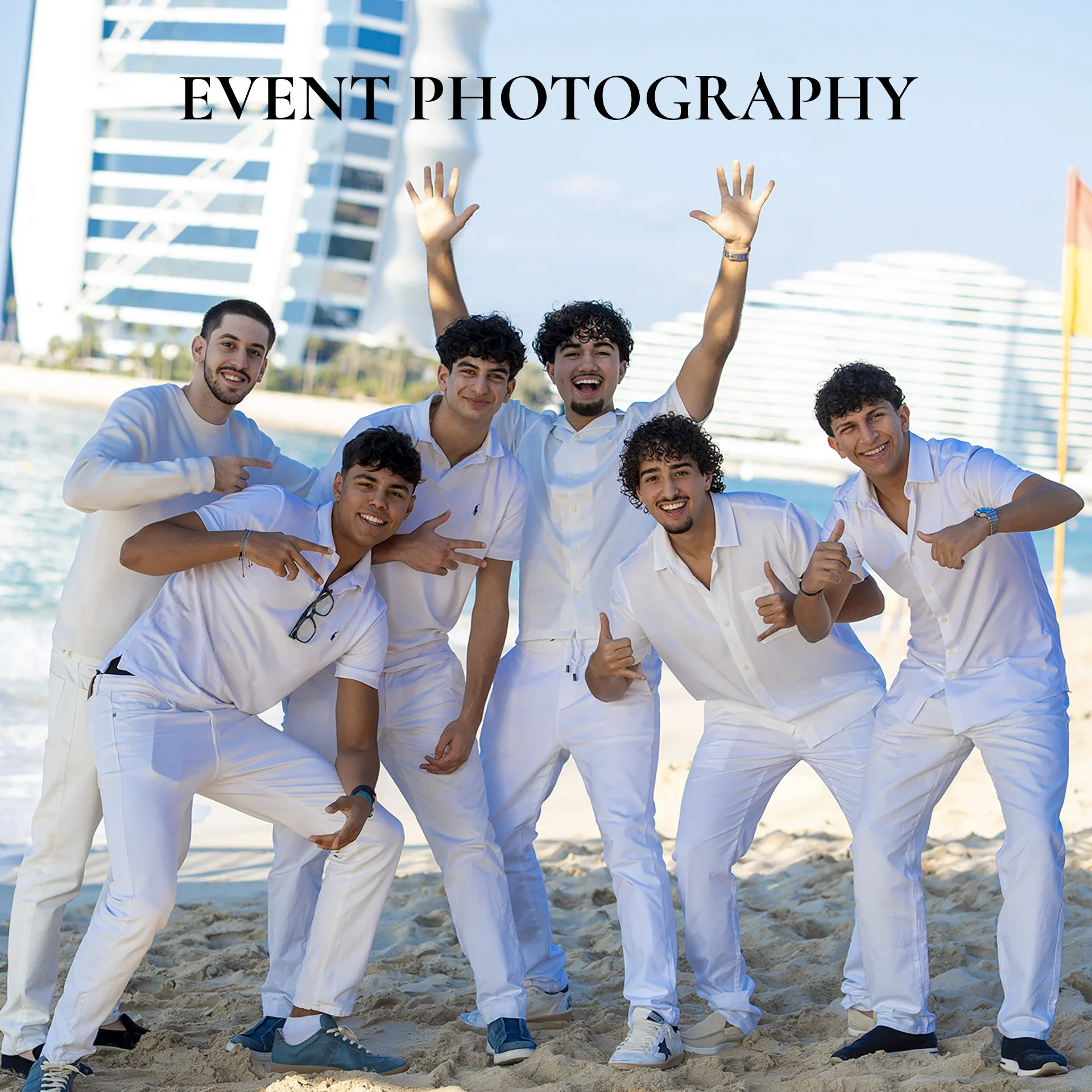 Group of six young men in white clothing posing and smiling on a beach with the Burj Al Arab hotel in Dubai in the background, during a sunny day.