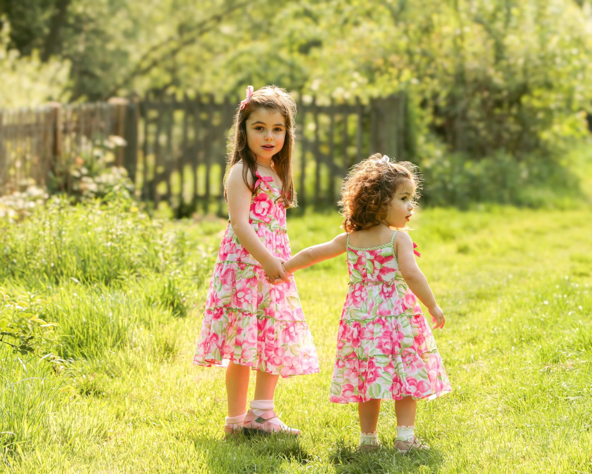 Two girls in matching pink floral dresses holding hands in a sunlit yard with green grass and trees.