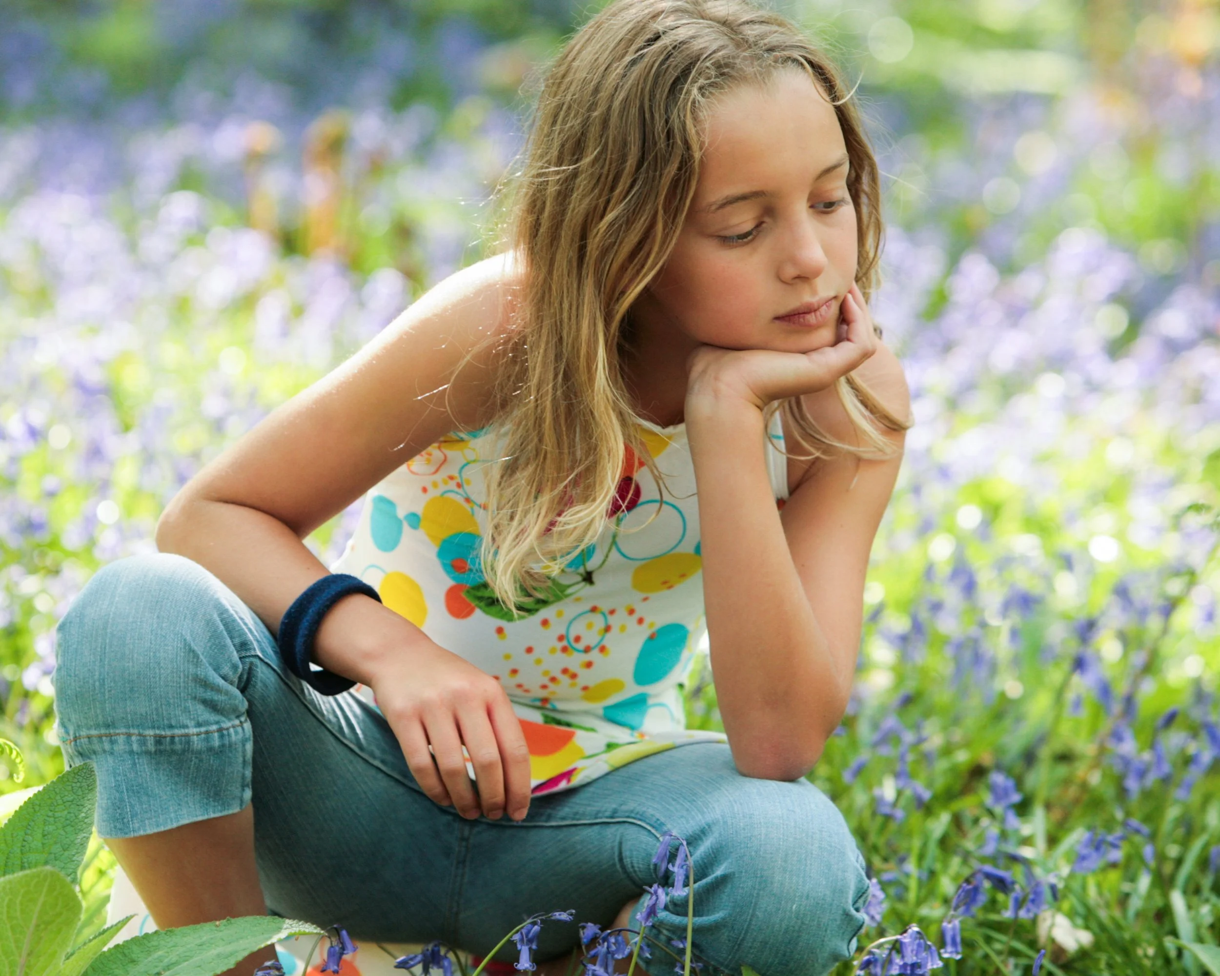 A young girl with long, wavy blonde hair sitting outdoors among purple flowers, resting her chin on her hand and looking pensively downward.