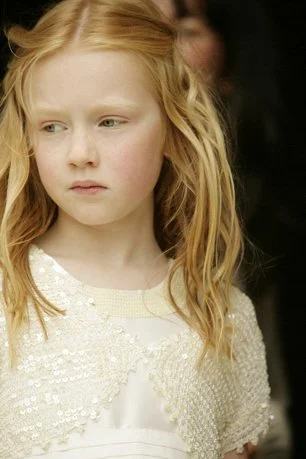 A young girl with red hair and a serious expression, wearing a white dress with pearl embellishments.