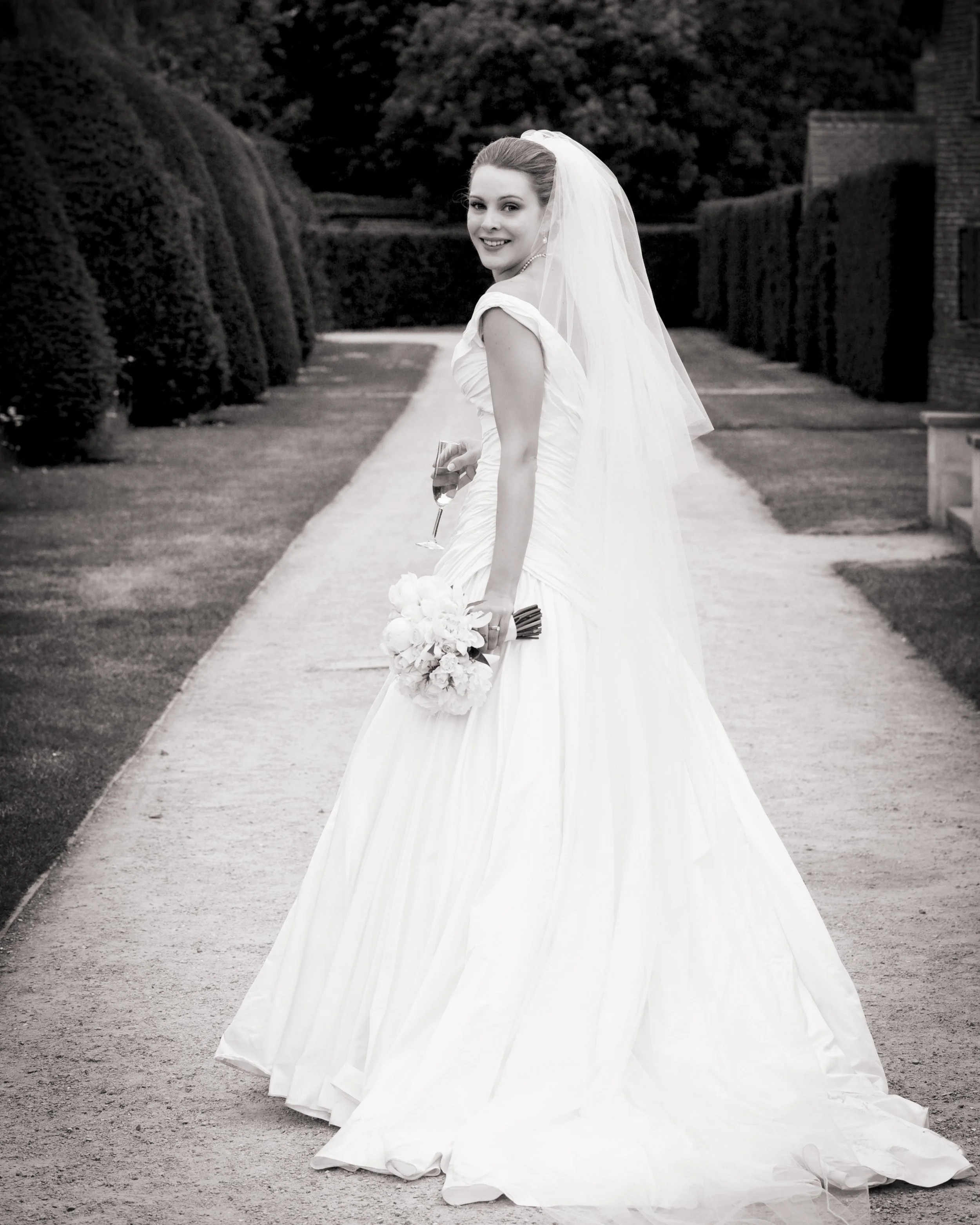 A smiling bride in a wedding dress holding a bouquet and a glass of champagne, standing on a garden pathway with hedges in the background.