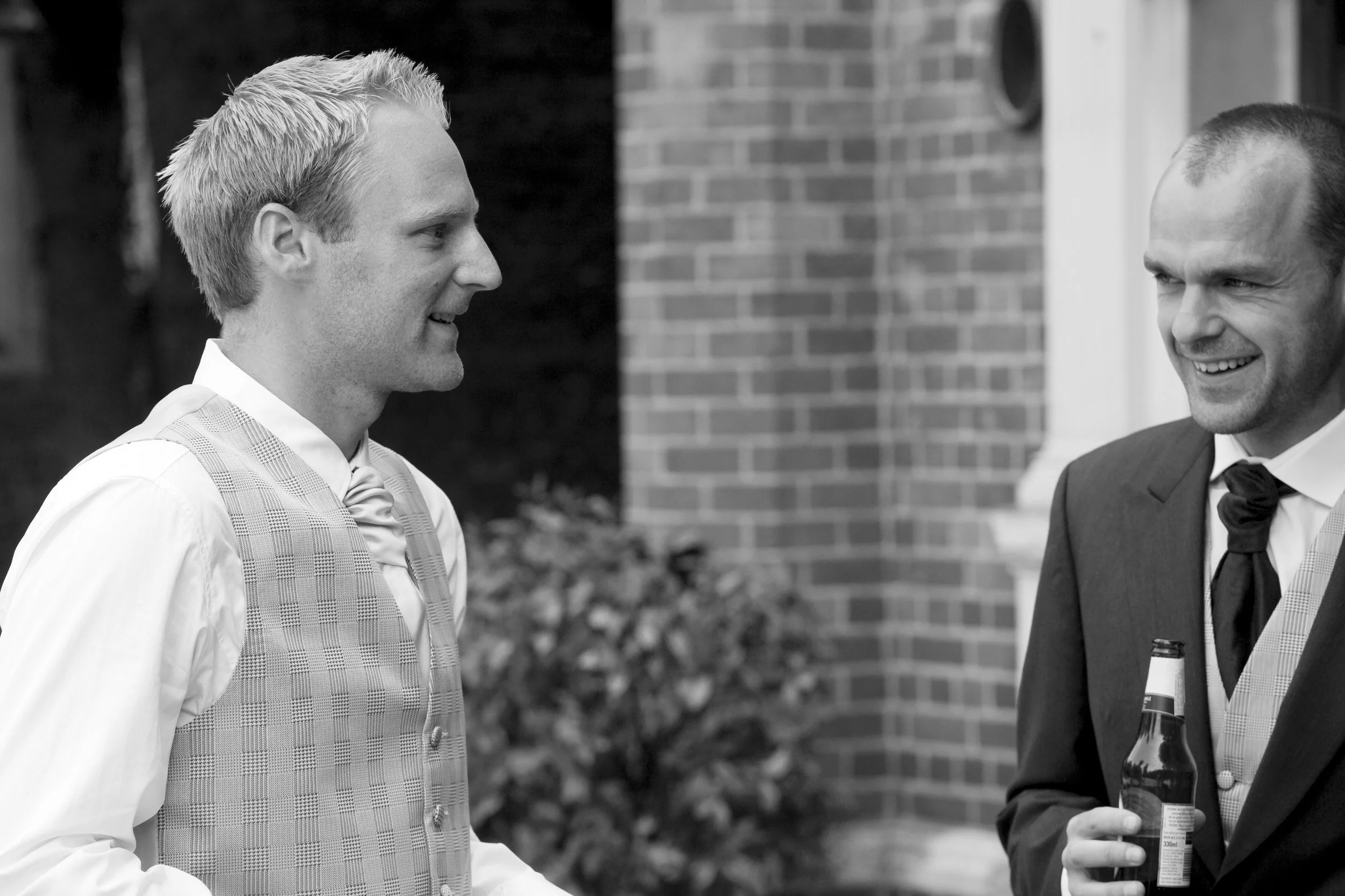 Two men in formal attire engaging in conversation outdoors, with one holding a beer bottle, smiling. Background includes brick wall and shrubbery.