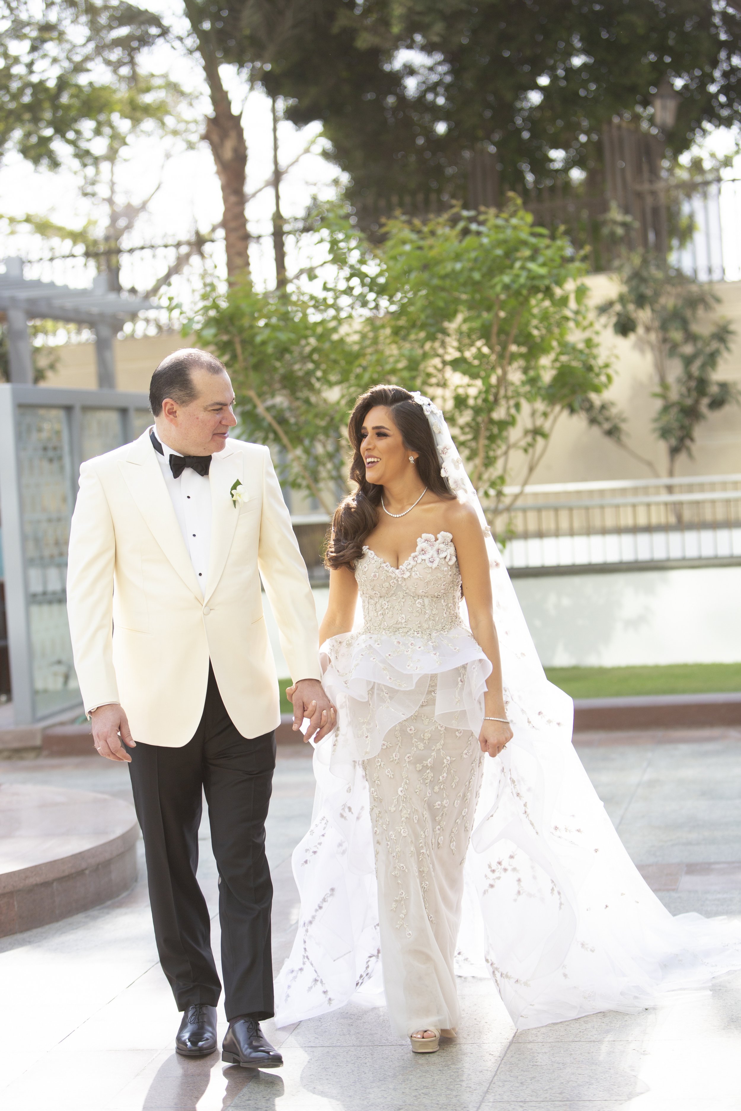 A bride and groom walking outside, holding hands, smiling at each other during their wedding day. The bride is in a detailed white wedding gown with a veil, and the groom is in a white tuxedo jacket and black pants.