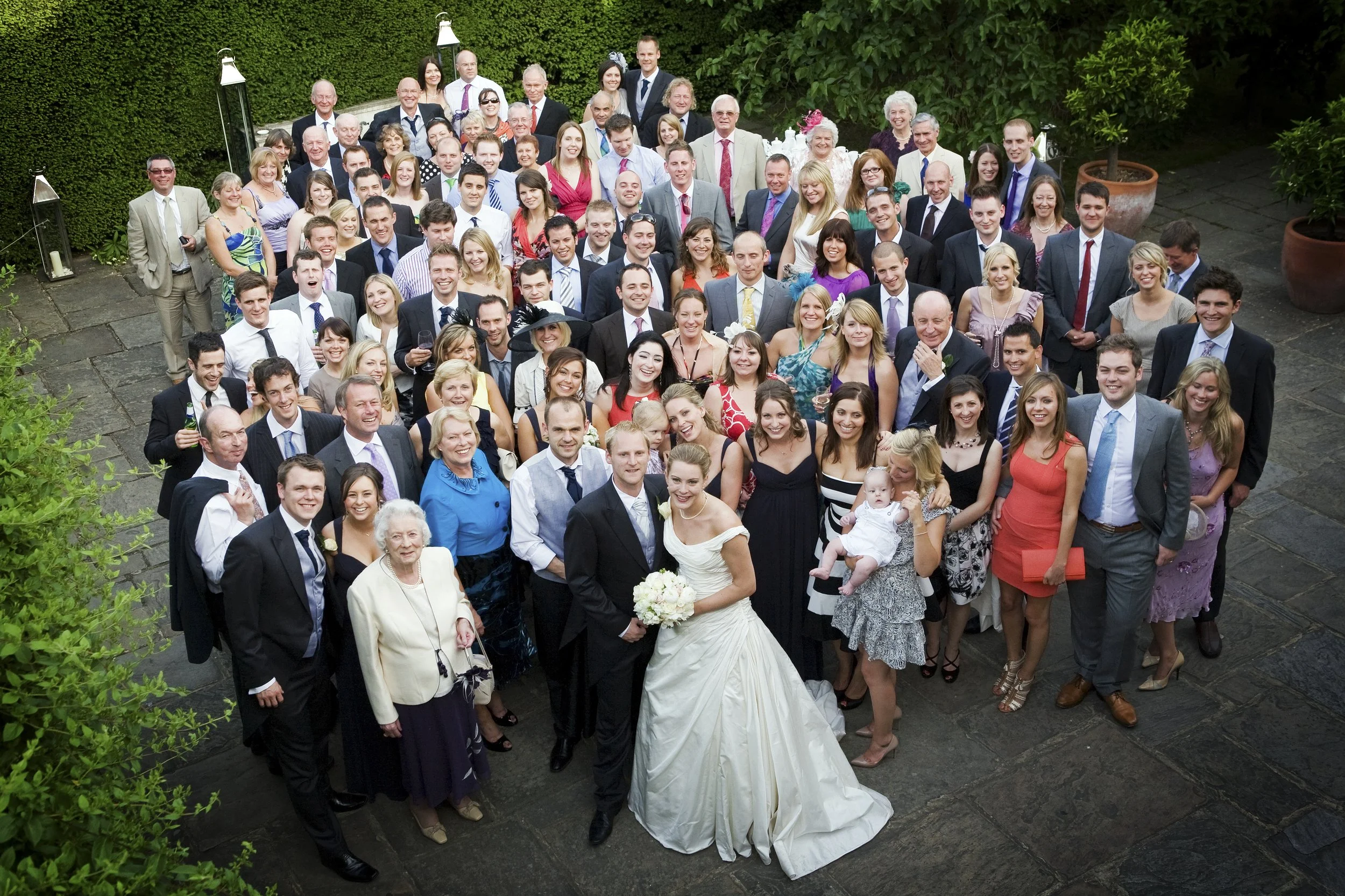 A large group of people, dressed in formal attire, gathered outdoors on a stone patio for a wedding celebration. The bride and groom are in the front, smiling and holding hands.
