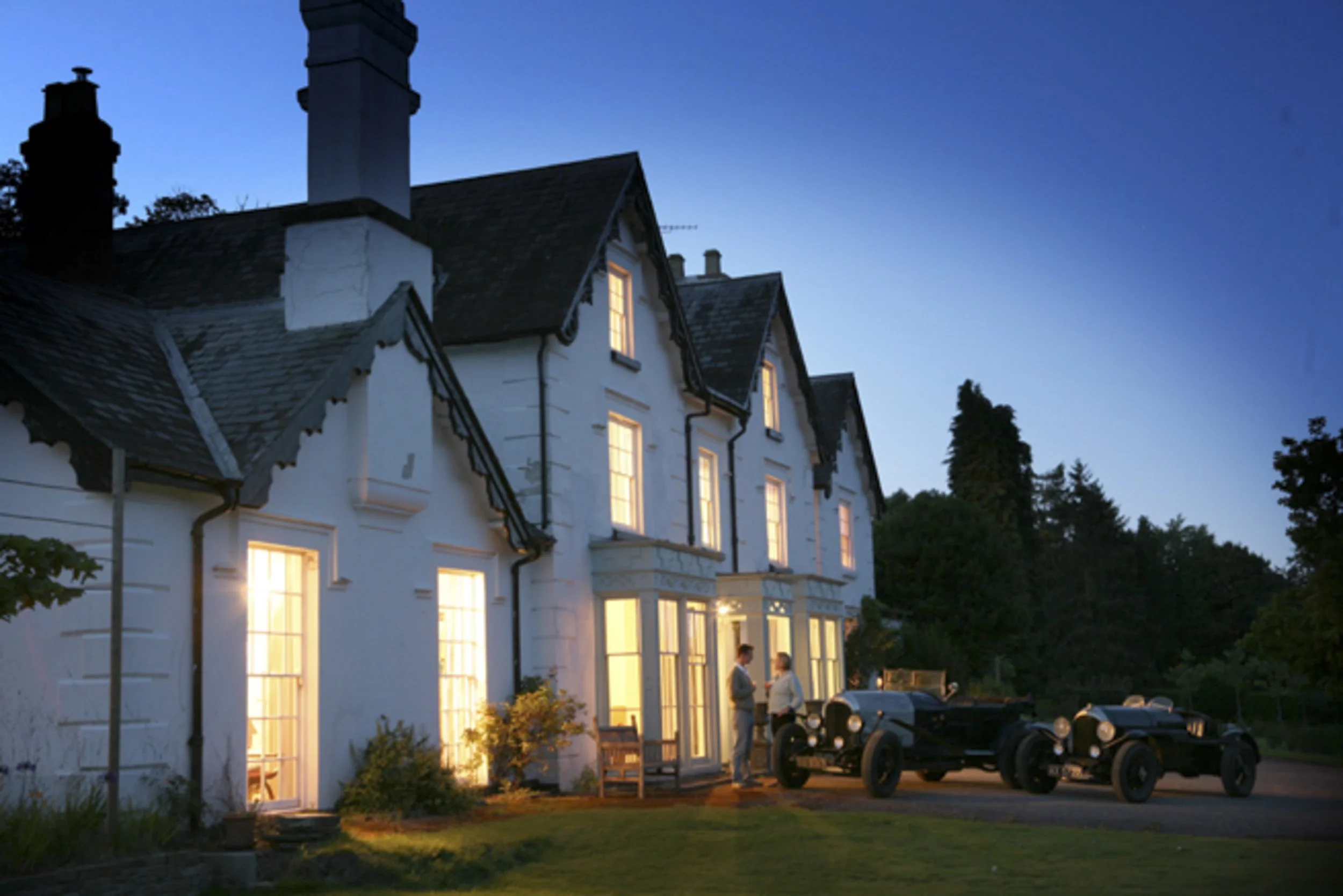 A white multi-story house with lit windows at dusk, two men talking outside near vintage cars, and a backdrop of trees and blue sky.