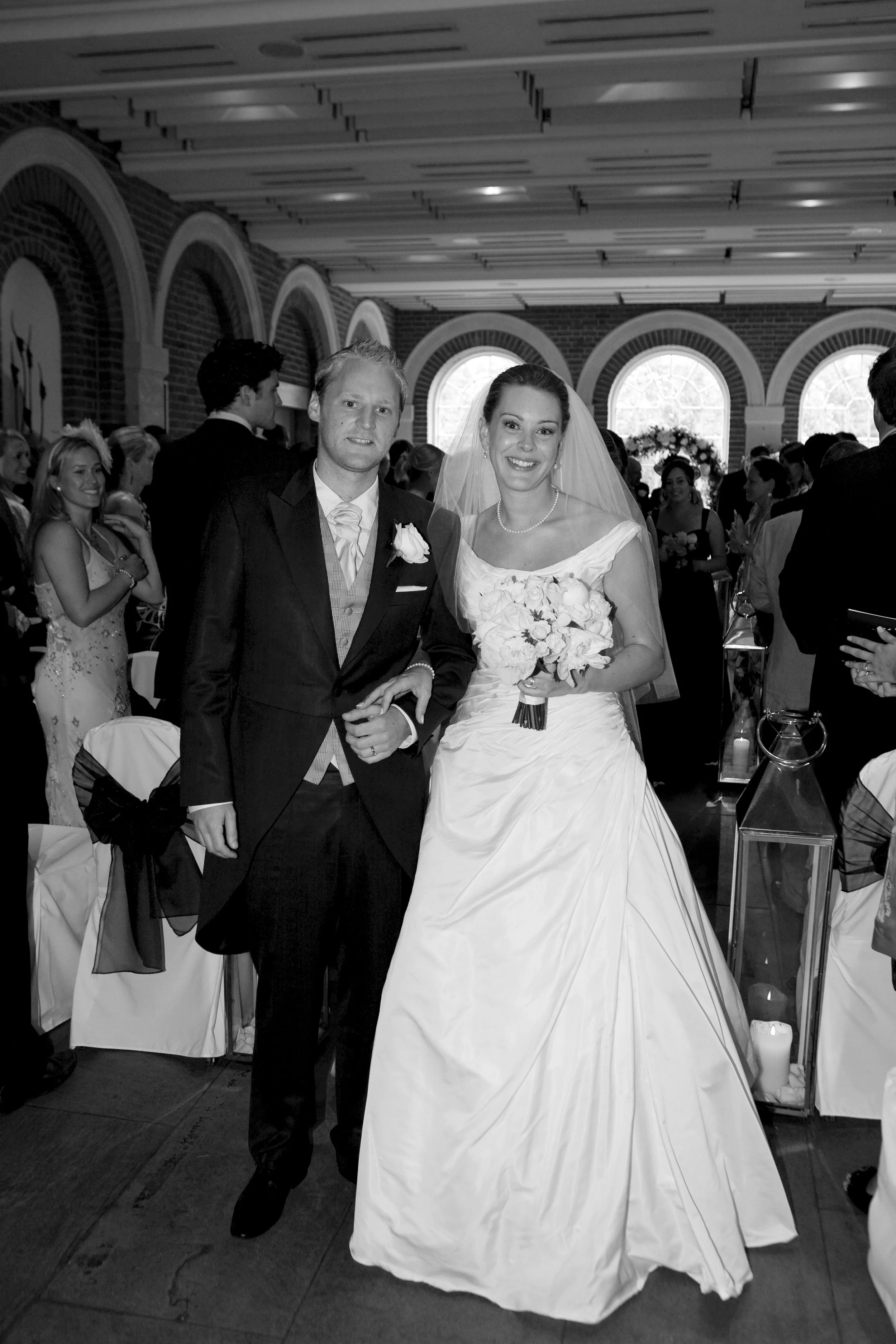 Black and white photo of a bride and groom at their wedding reception, smiling and standing close together in a decorated venue with arched windows and candles.