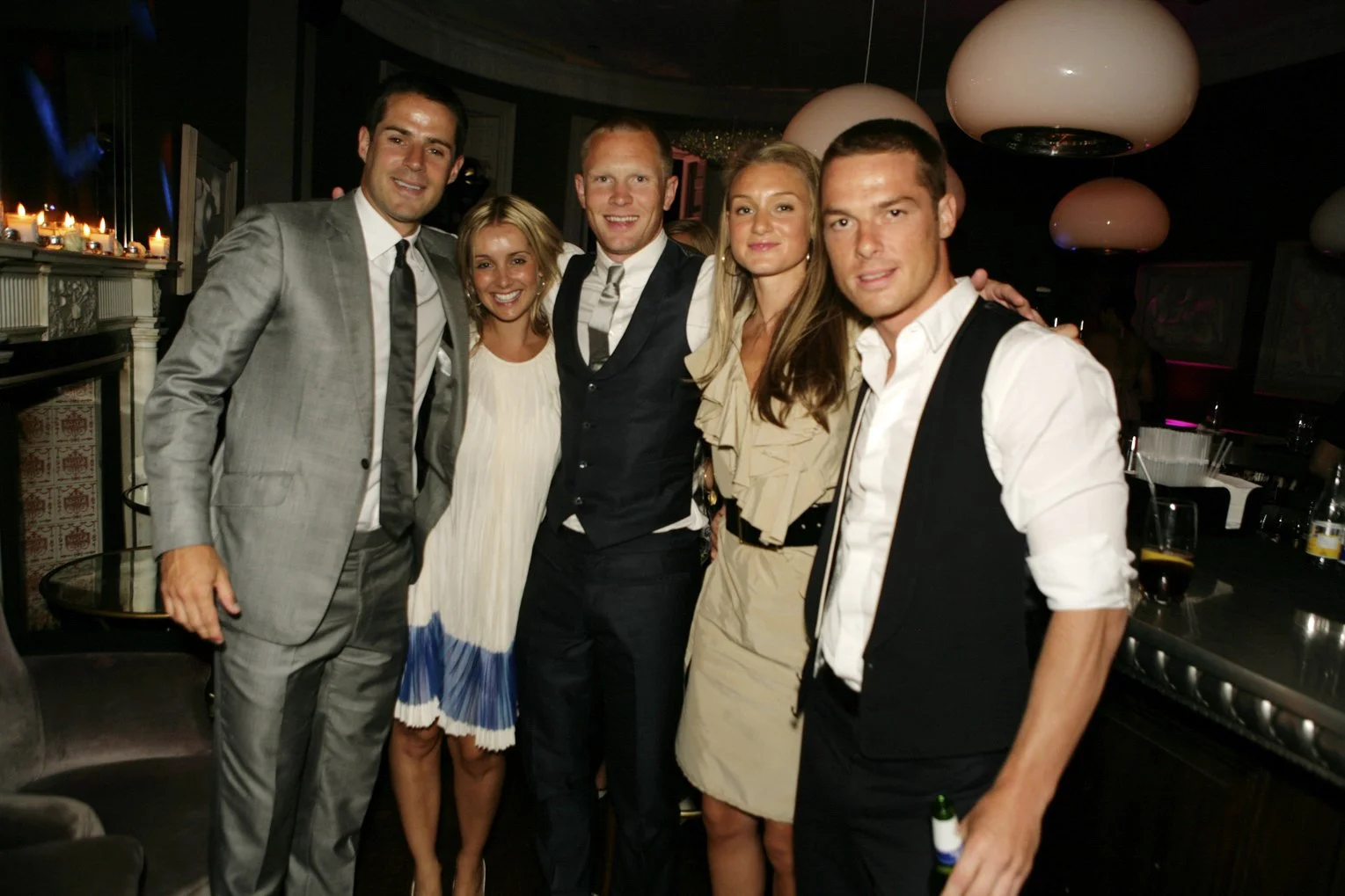 Group of five adults dressed in formal and semi-formal attire, smiling and standing together indoors at a social event or party.