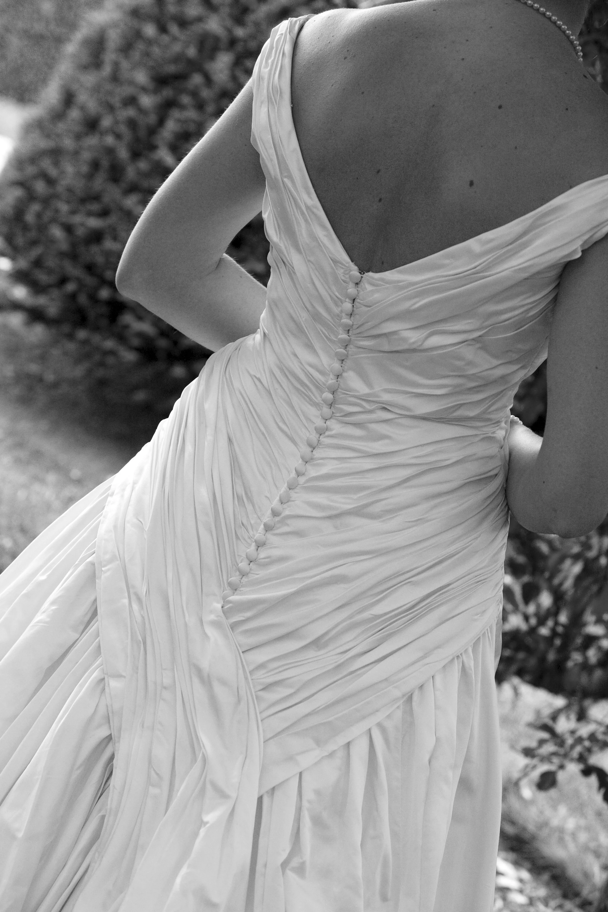 A black and white photo of a woman in a wedding dress, showing the back details of the dress with buttons, outdoor setting in the background.