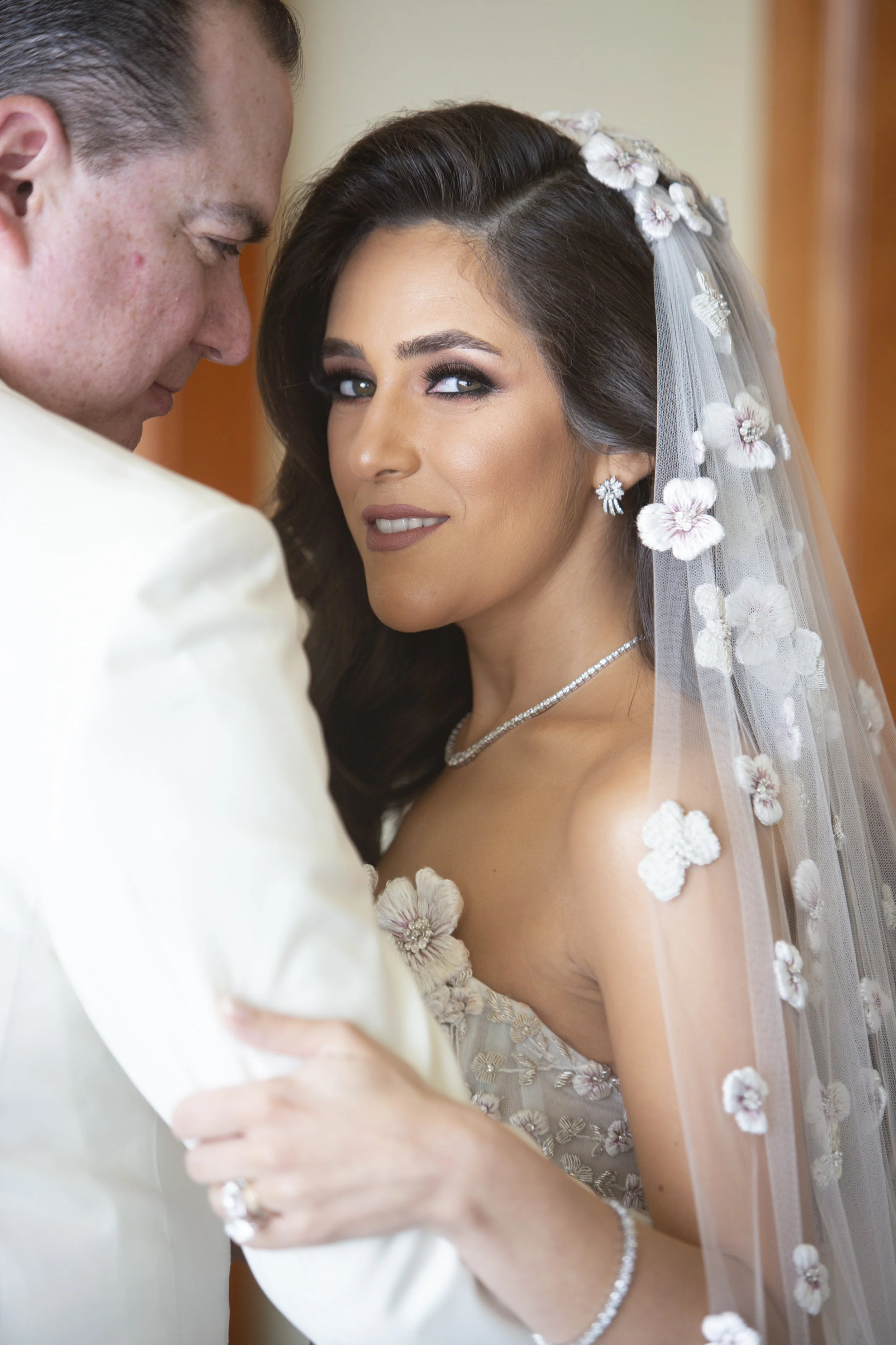 Bridal portrait of a bride with dark hair, wearing a floral wedding dress, veil, diamond jewelry, and makeup, being embraced by a groom in a white suit.