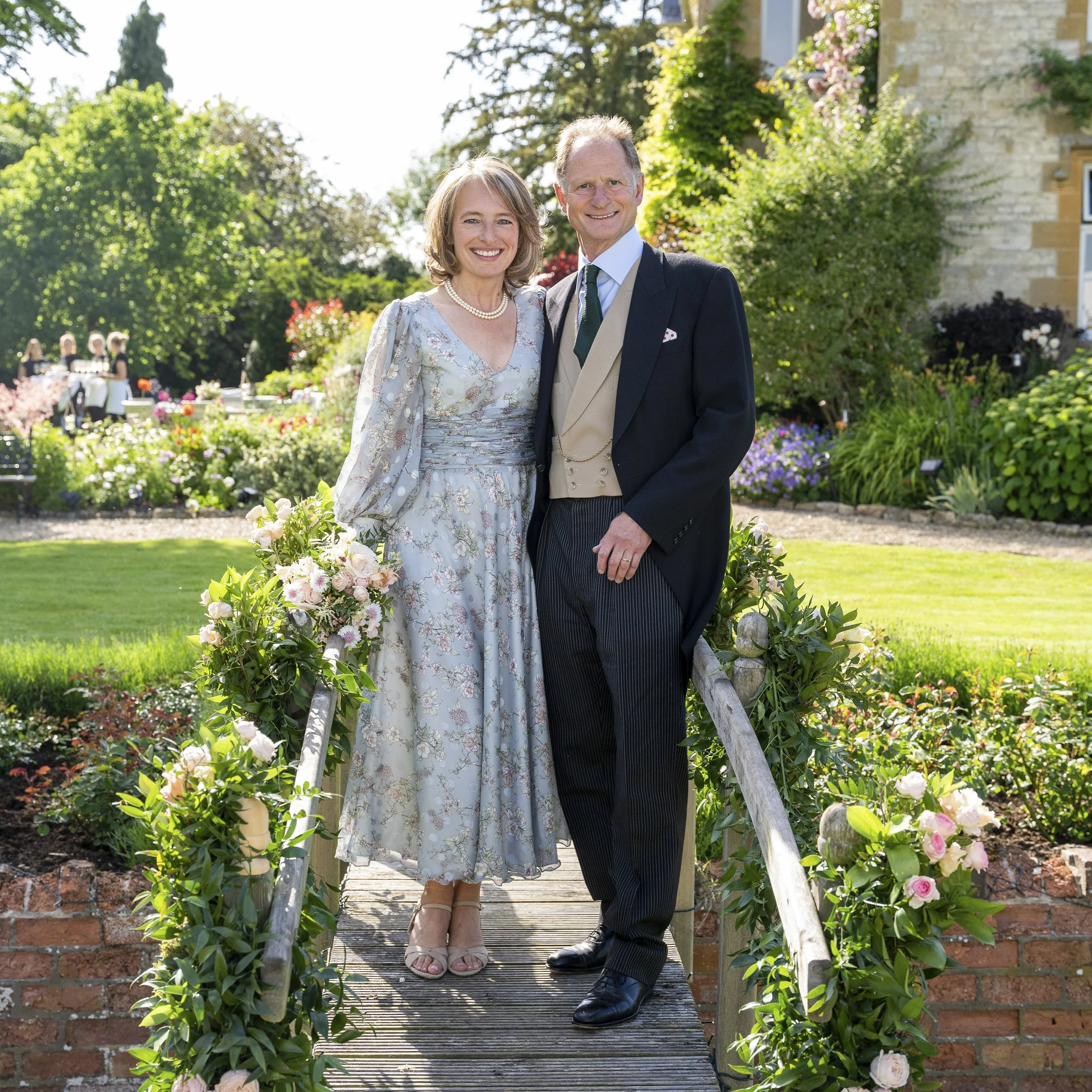 A smiling woman and man in vintage formal attire standing on a small wooden bridge decorated with flowers, in a lush garden setting during daytime.