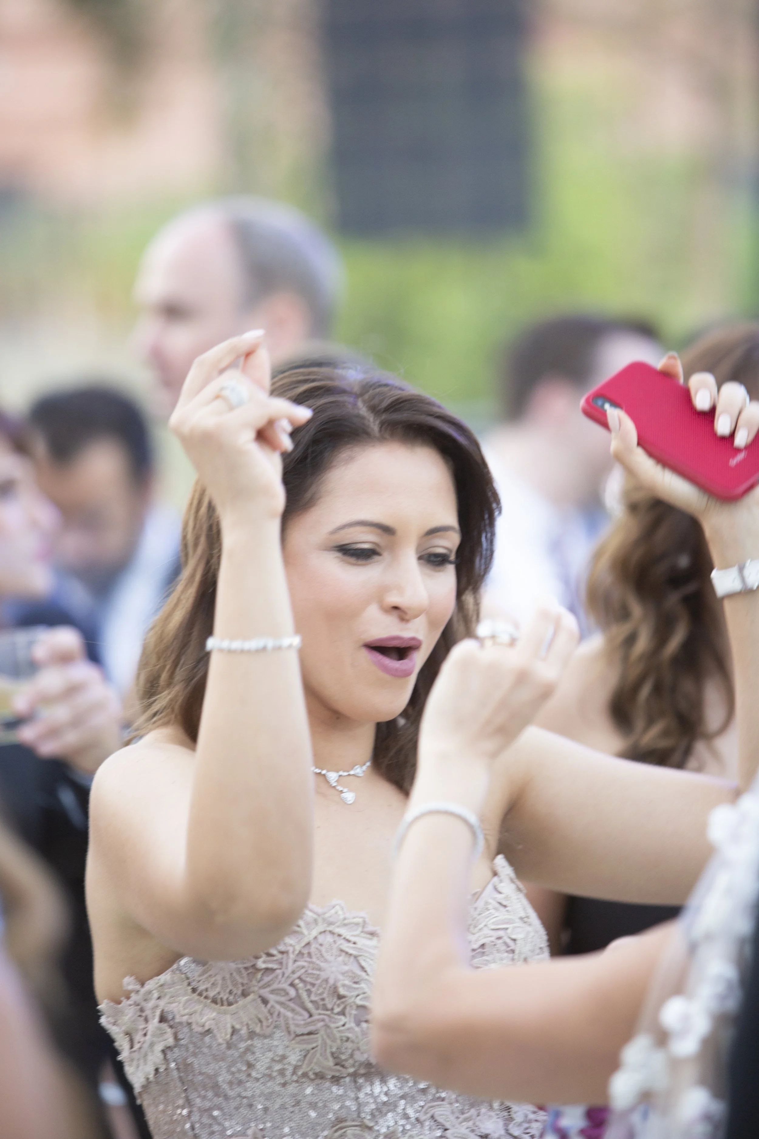 A woman with brown hair wearing a light-colored lace dress dancing at an outdoor event, with other people and trees blurred in the background.