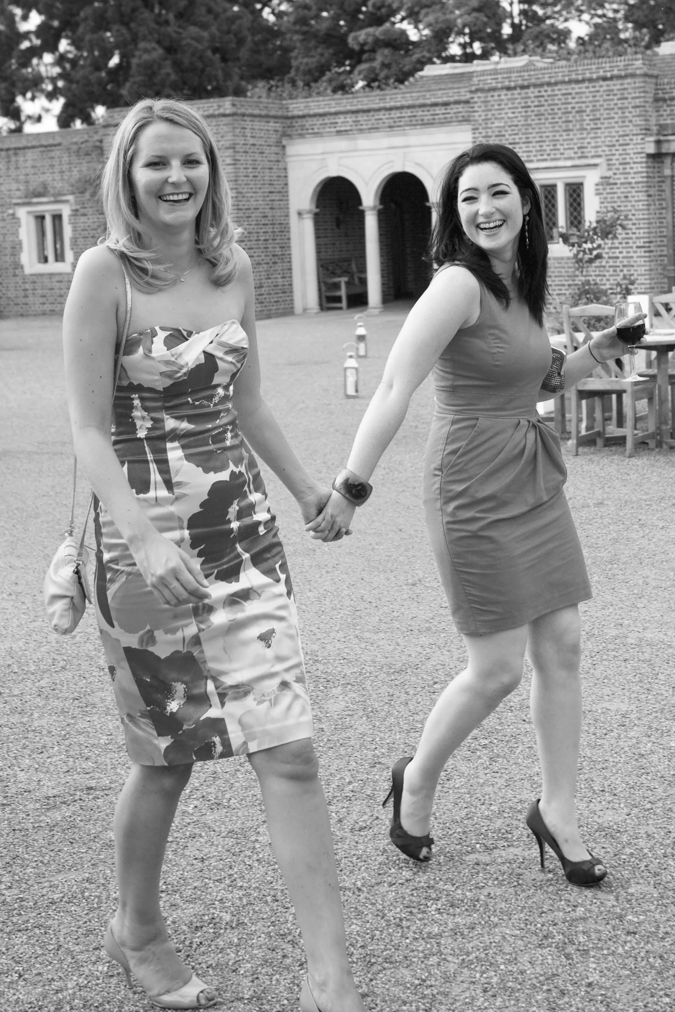 Two women holding hands and walking outdoors, smiling. One is holding a glass of wine, wearing a floral dress. The other is wearing a plain dress and high heels, holding a glass of wine.