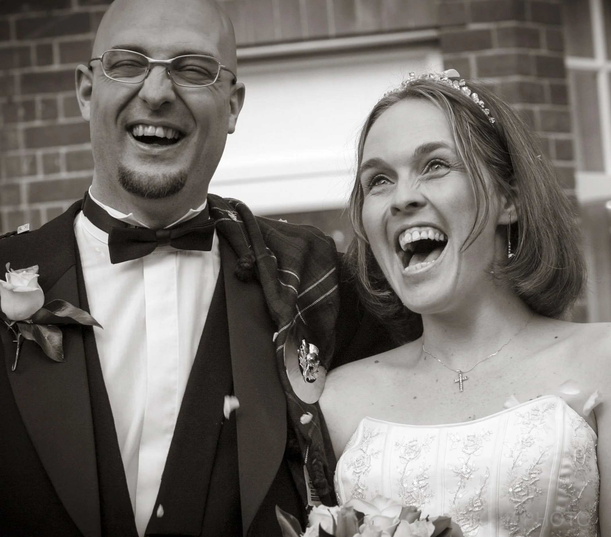 A happy bride and groom laughing together at their wedding, standing outside near a brick wall.