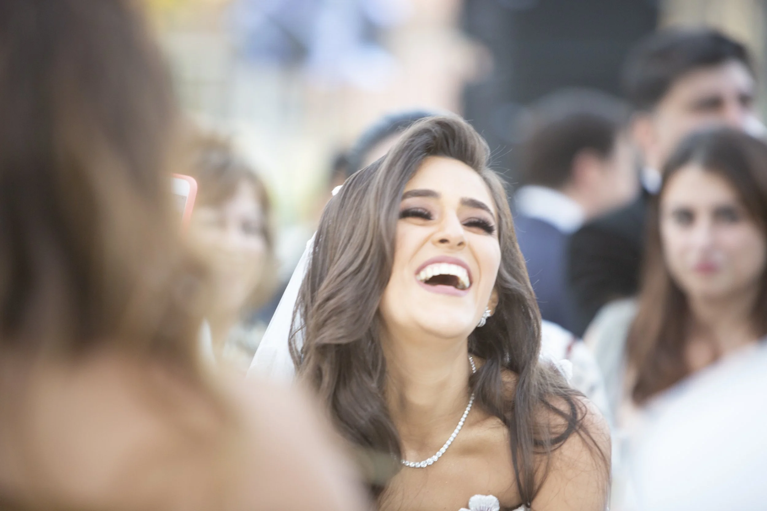 A woman with long dark hair, wearing makeup and jewelry, laughing at a wedding reception surrounded by other people.