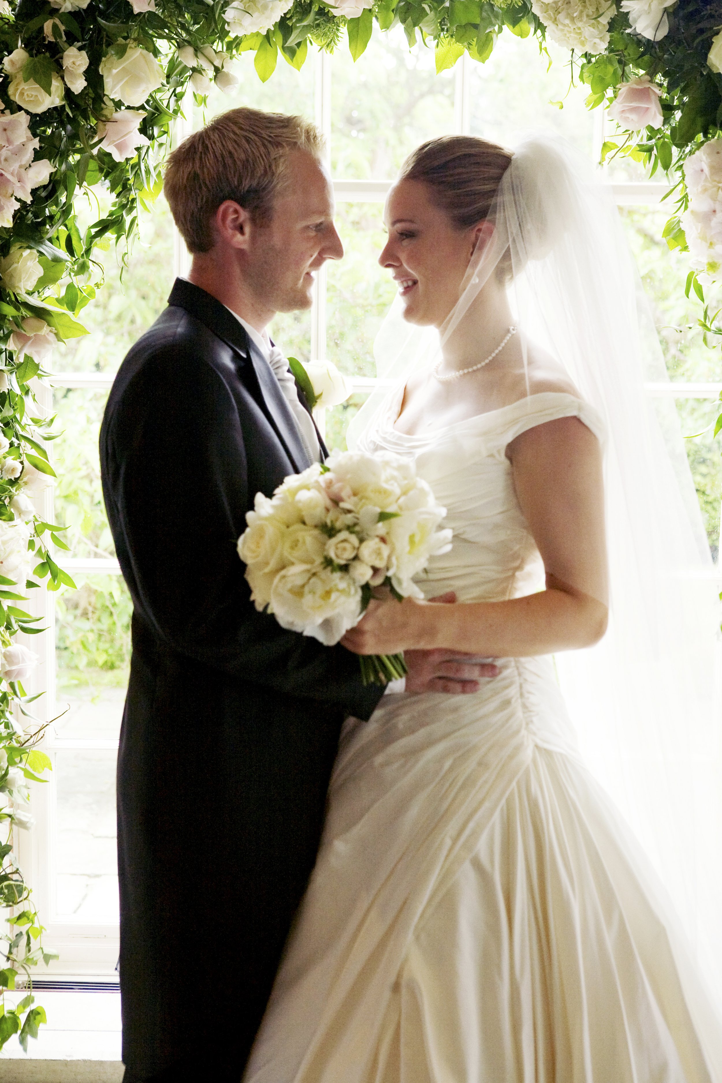 A bride and groom standing face to face under a floral arch, smiling at each other. The bride is holding a bouquet of white roses and wearing a white wedding gown with a veil, while the groom is in a black suit with a white shirt and tie.