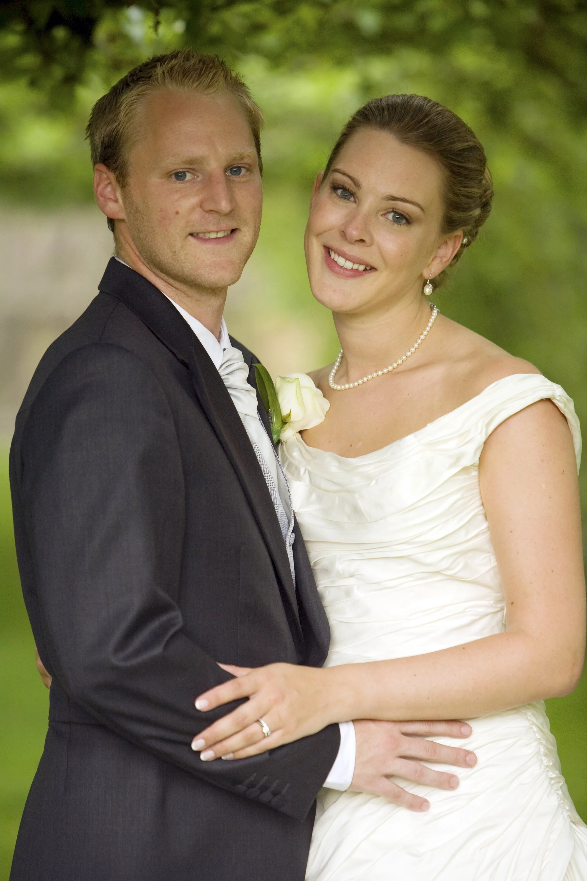 A newlywed couple, a man in a dark suit with a white shirt and tie, and a woman in a white wedding gown with pearl jewelry, standing outdoors with green foliage in the background, smiling and holding hands.