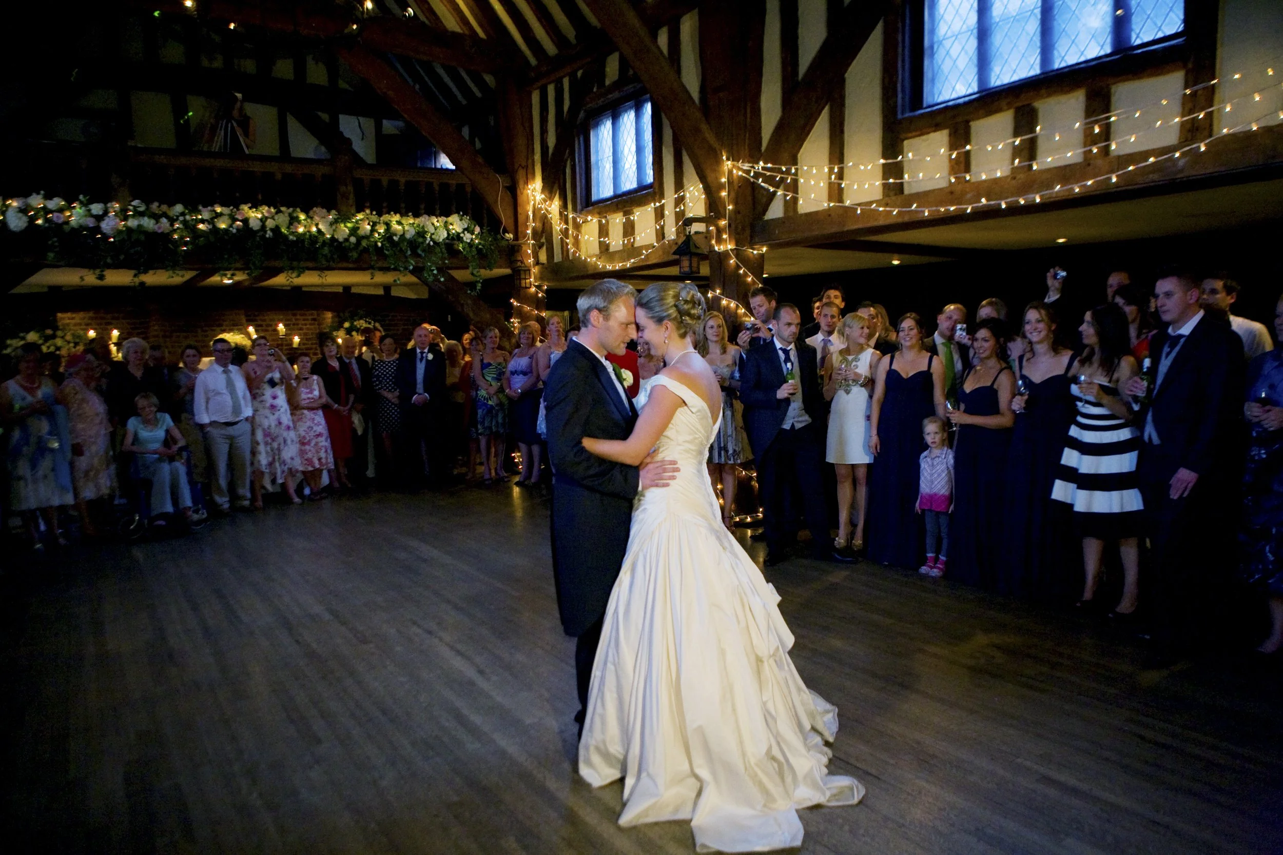A bride and groom sharing their first dance at a wedding reception with wedding guests surrounding them in a decorated, rustic-style venue.