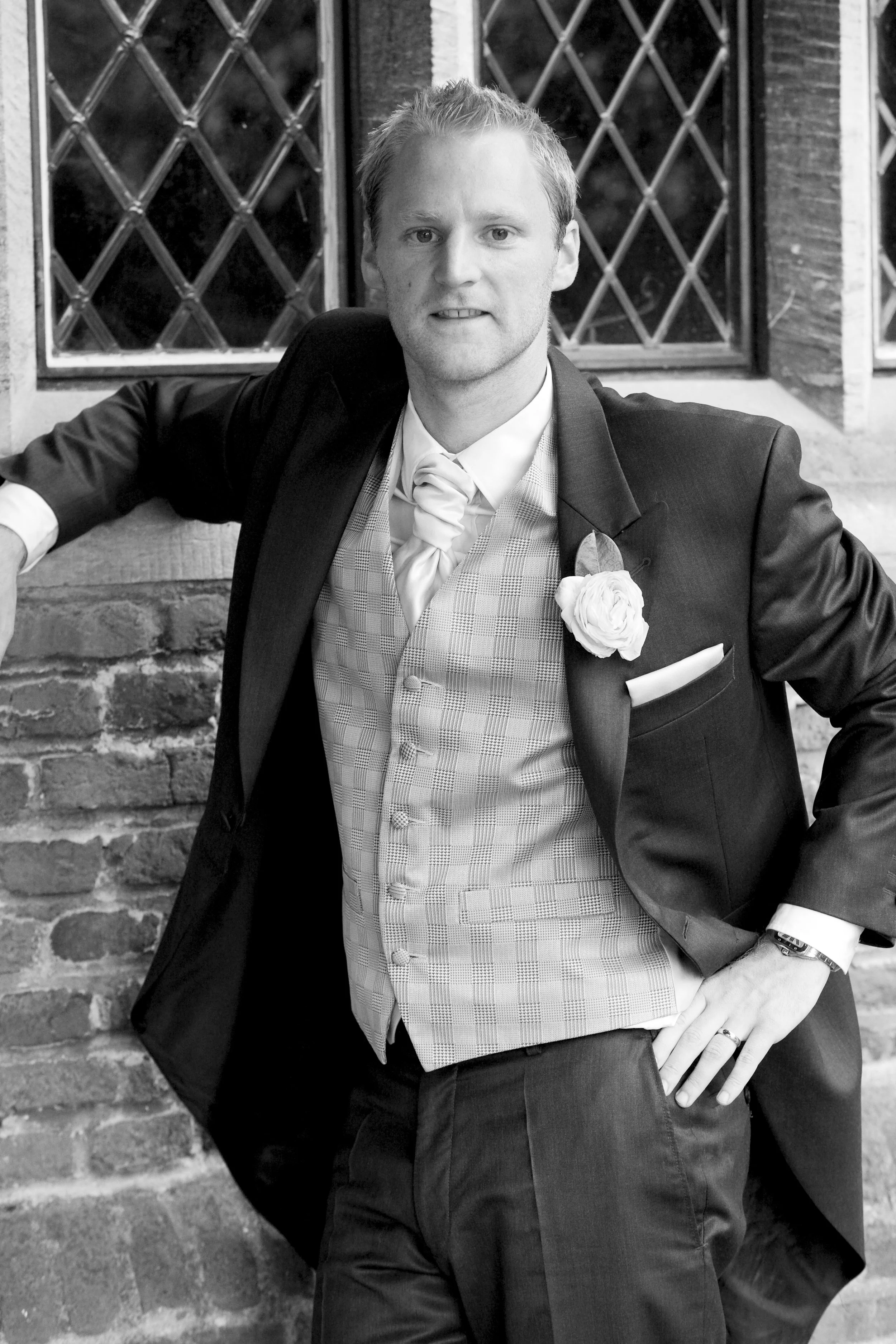 A man in a tuxedo with a boutonniere, standing outside against a brick wall with diamond-pane windows.